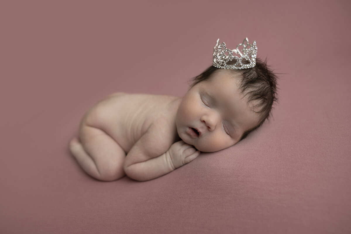 A newborn baby lays asleep on a pink blanket wearing a silver crown.