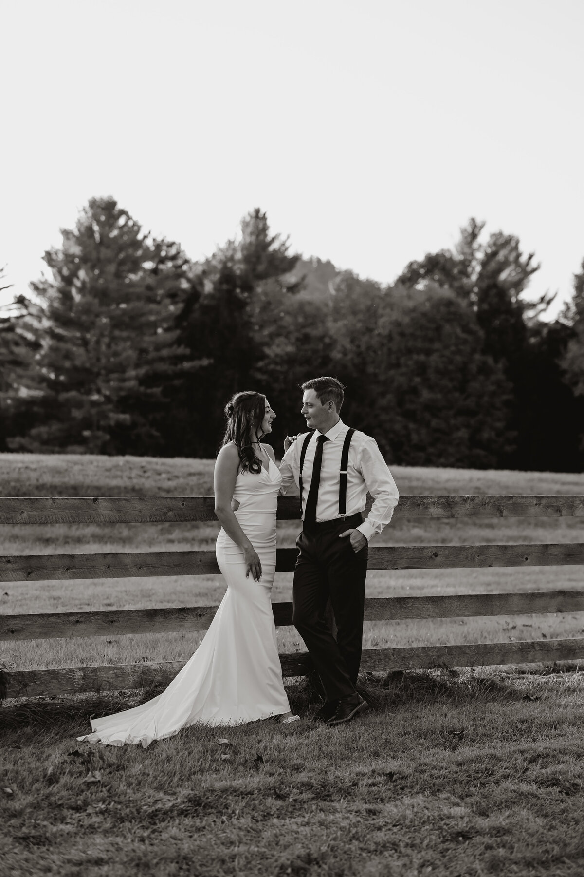 In a timeless black and white frame, a bride in a flowing white dress and her groom in suspenders stand by a wooden fence beneath towering trees. Their smiles illuminate the rural setting, captured masterfully by an elopement photography expert from New England.