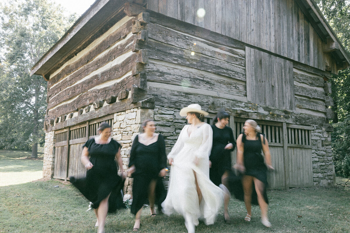 Bridal party features motion as they walk away from a barn and towards a sunlit meadow. 
