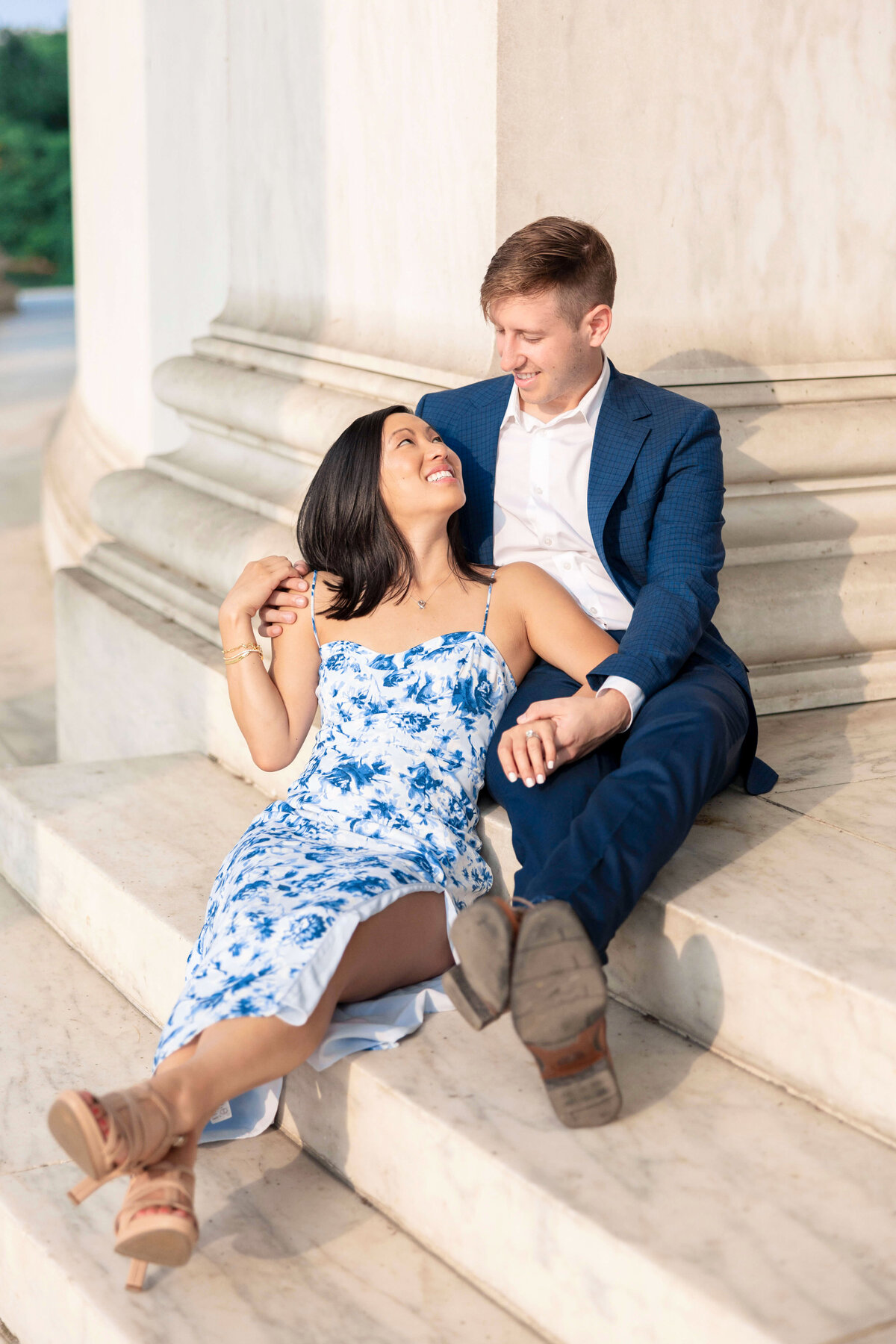 A couple is smiling and sitting on marble steps. The woman is wearing a blue and white floral dress, and the man is in a blue suit. They are sitting close, holding hands, and looking at each other affectionately.