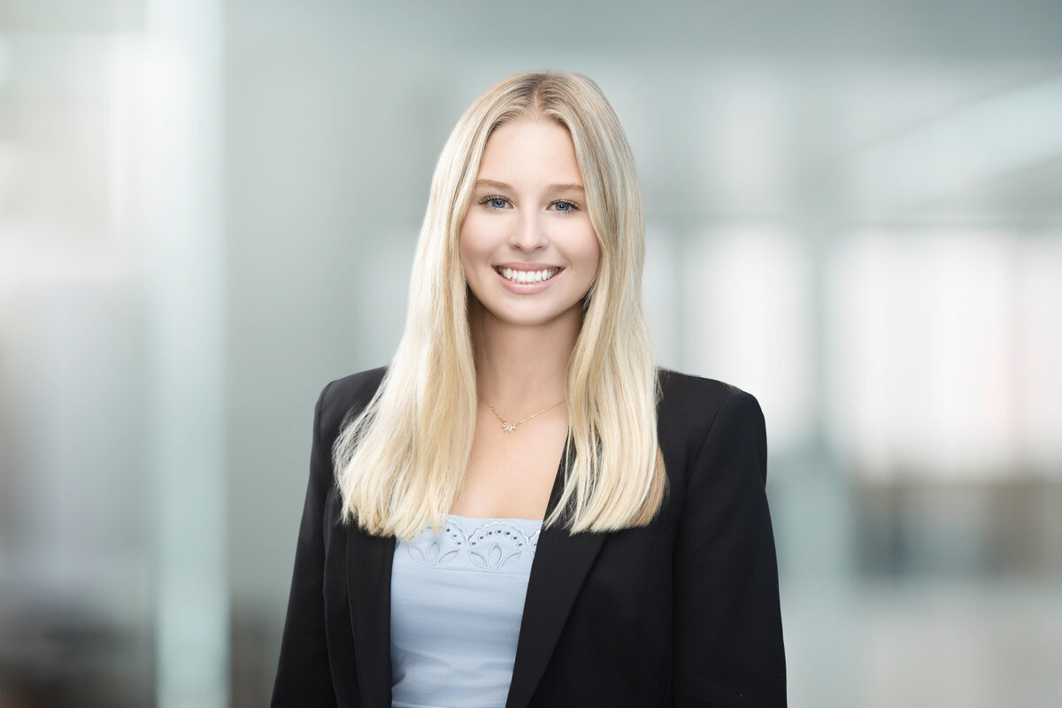 Professional headshot of a young woman from Messer Construction, smiling confidently in a black blazer and light blue top, with straight blonde hair, photographed at Janel Lee Photography studio in downtown Cincinnati, Ohio. She stands against a bright, modern, blurred office backdrop that complements her approachable and professional expression.
