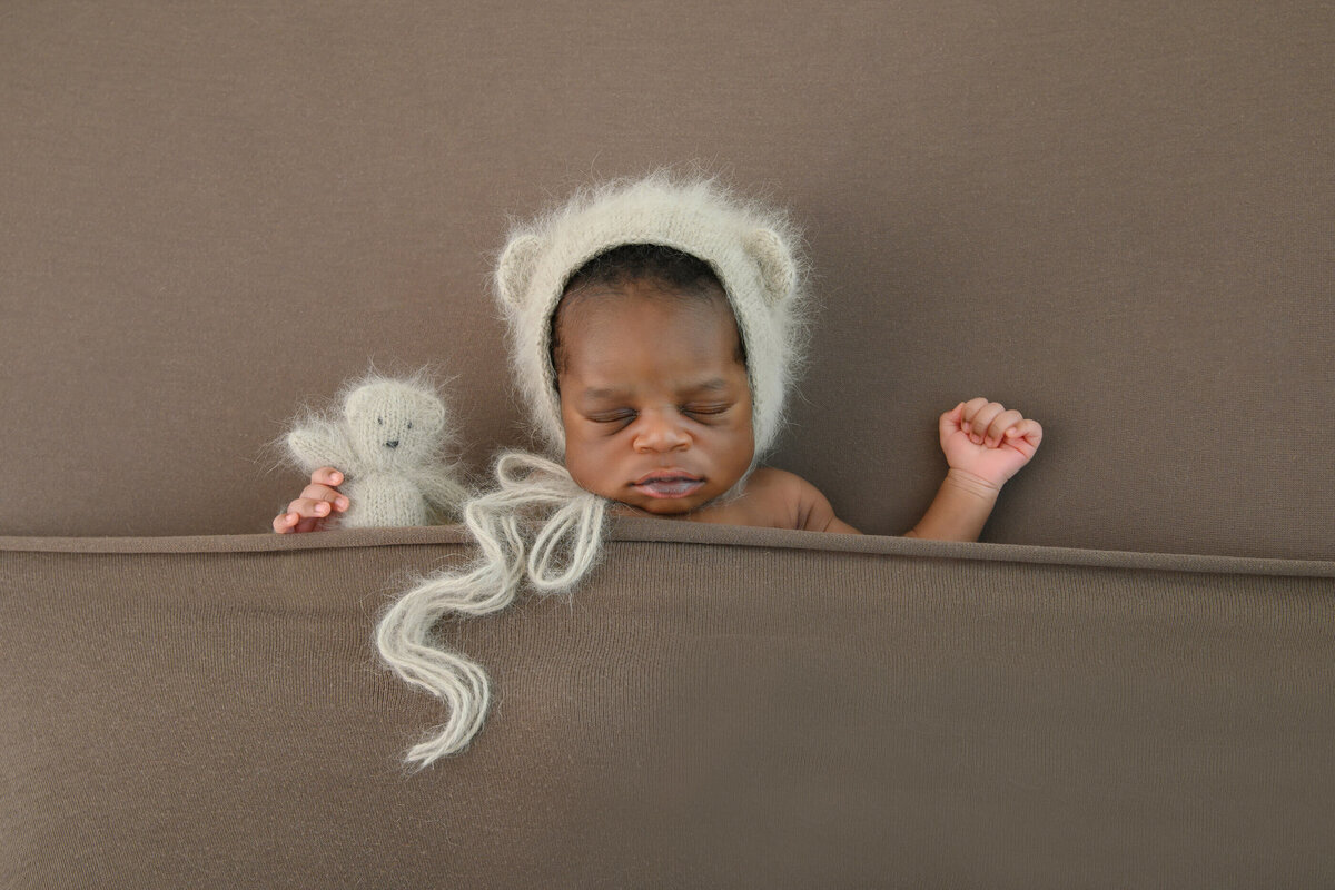 Newborn baby boy wearing a beige bear hat and holding a tiny beary on a brown background