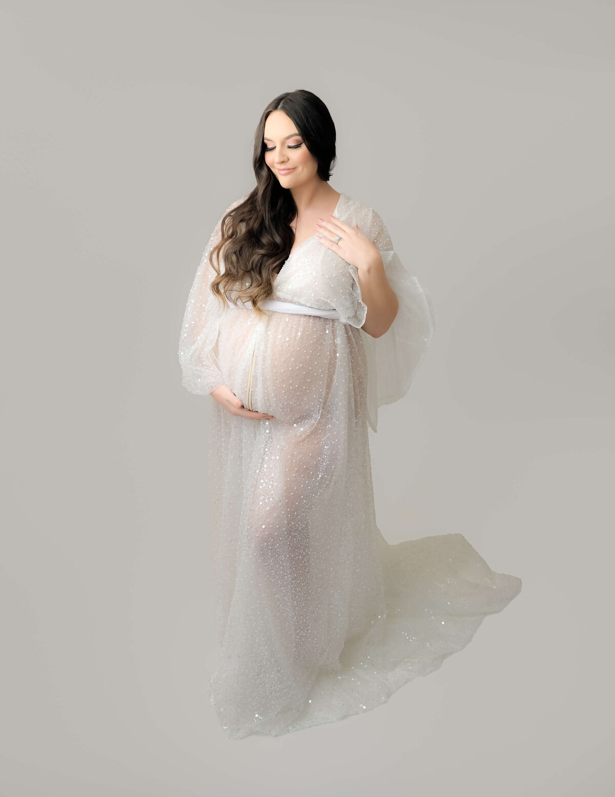 Beth Benecki captures this full length portrait of an expecting mother during a maternity photo session in Bremerton. Against a white background, she is in a white sequin sheer robe.  Her long brown hair rests over one shoulder as she looks down at her baby bump. Image photographed by Beth Benecki.