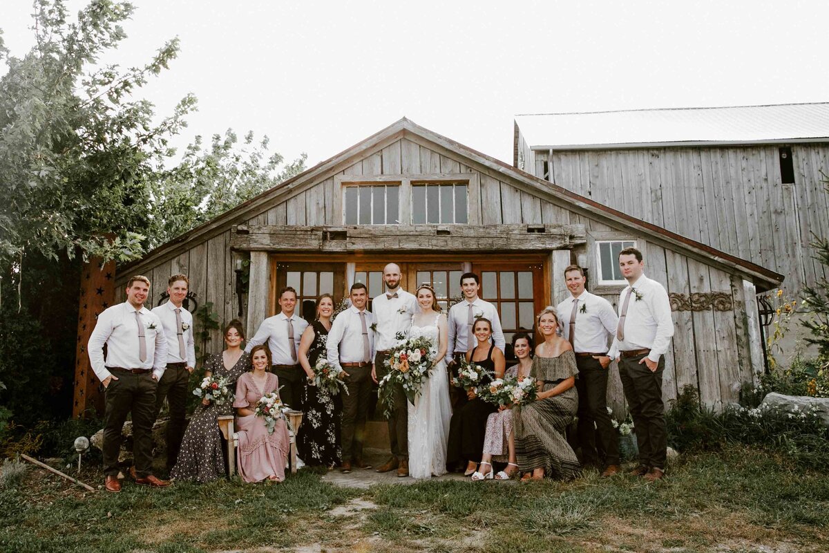 Bride, groom, and wedding party posing in front of rustic barn for formal wedding day portrait.