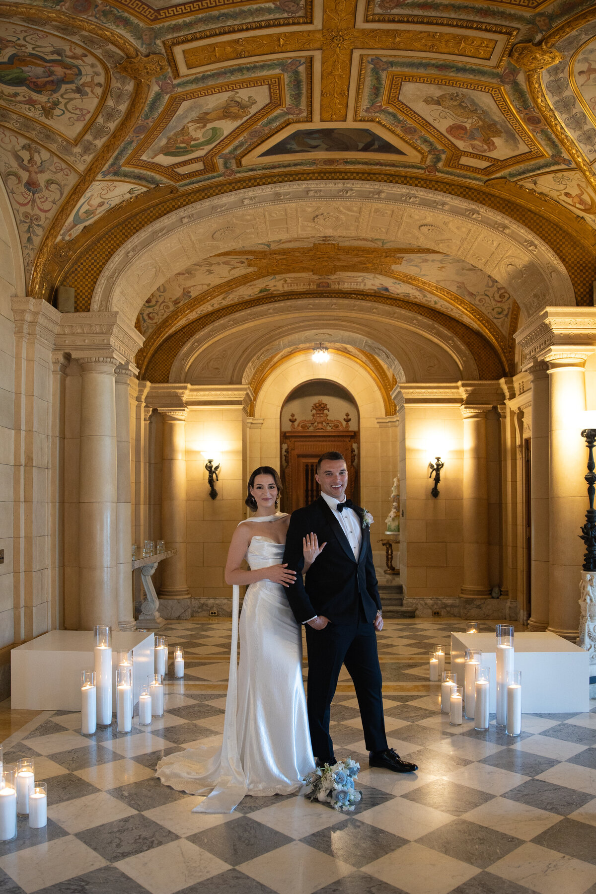 Aldrich Mansion Wedding Photos | Kelsey Sheehan Photography Timeless Rhode Island Weddings | A couple in formal attire stands in an elegant hallway with tall, arched ceilings. Lit candles line the floor, creating a romantic ambiance.