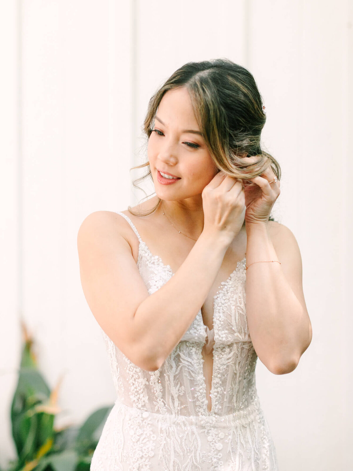 A bride in a lace wedding dress adjusts her earring with a gentle smile. Soft lighting and a serene background.