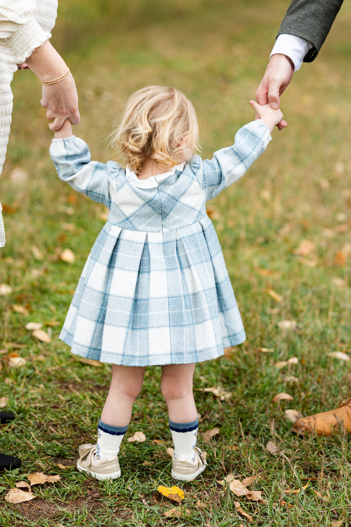 Photo of a toddler girl in a blue and white plaid dress walking away from the camera while holding her mom and dad's hands.