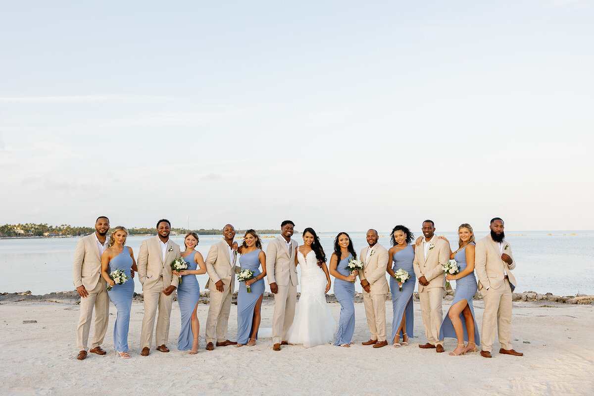 Andressa and Jonathan pose with their bridal party in chic beige suits and blue dresses, standing along the sandy shore of Table Key, Islamorada. Photography by Claudia Amalia Wedding Photography.