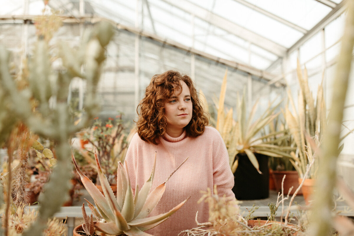 High school senior with short hair wearing a pink sweater posing in a greenhouse