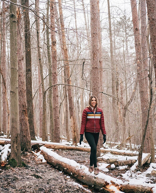 A woman in a red jacket and black pants walks through the woods, showcasing Cotopaxi's outdoor gear.