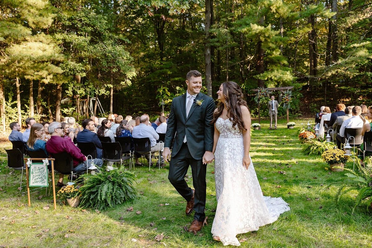 A couple leaves their ceremony hand in hand officially married. They are looking at one another and smiling. 