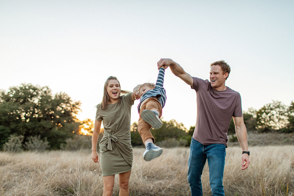 Family swinging their child in a field as they laugh, photographed by Sara James in San Antonio.