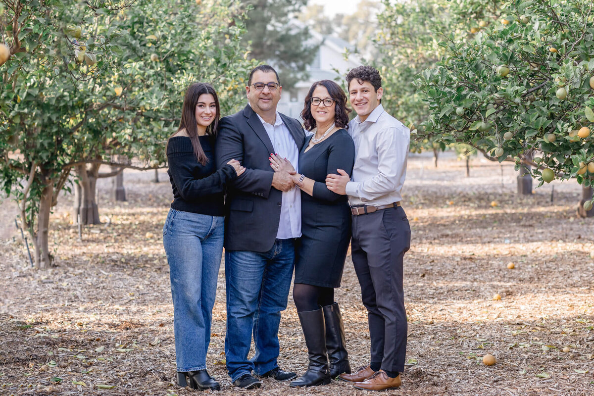 A family of four wearing black and grey standing with their arms around each other between two rows of green lemon trees, smiling at the camera.
