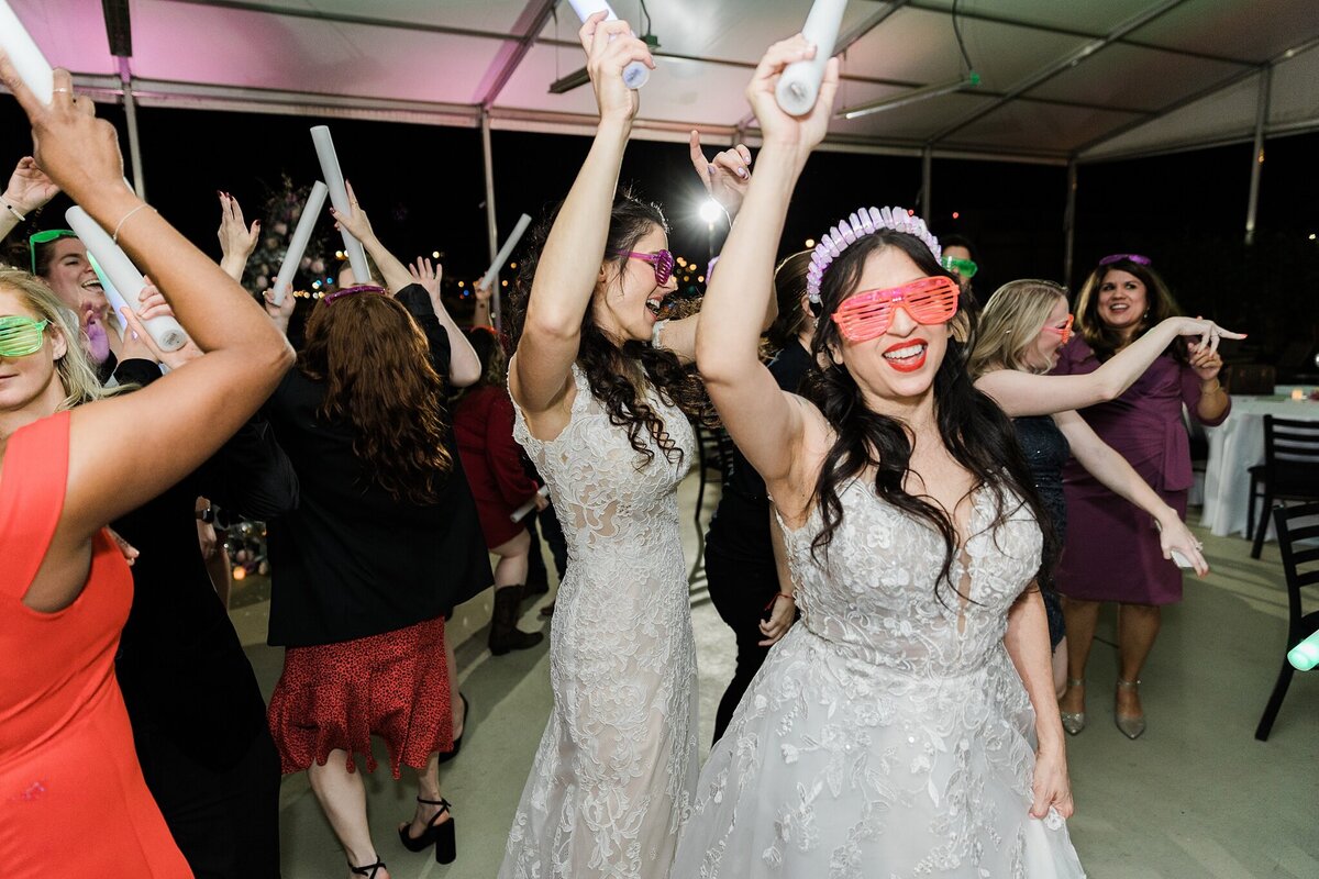 Two brides partying hard and dancing with their wedding guests during their wedding reception in Dallas, Texas. The bride on right is wearing a short sleeve, long, intricate white dress, fun sunglasses with slits, and a light up crown. The bride on the left is wearing a short sleeve, shorter, intricate, white dress with similar sunglasses. The brides and most of the guests are all waving light up foam sticks around with dancing.