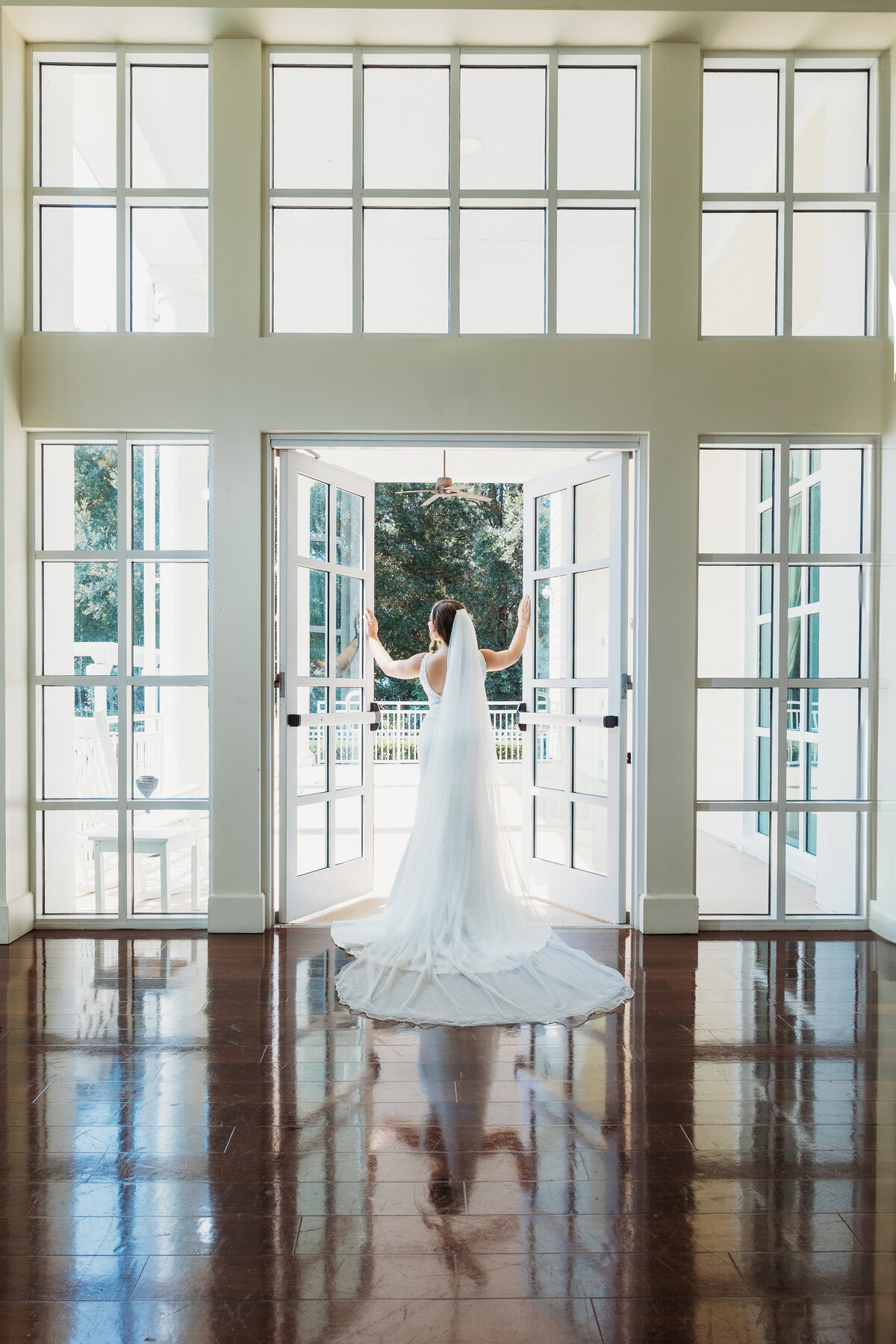 Bride stands in sunlit doorway of Lake Mary Event Center, her long veil and gown train dramatically fanned behind her on polished floors.