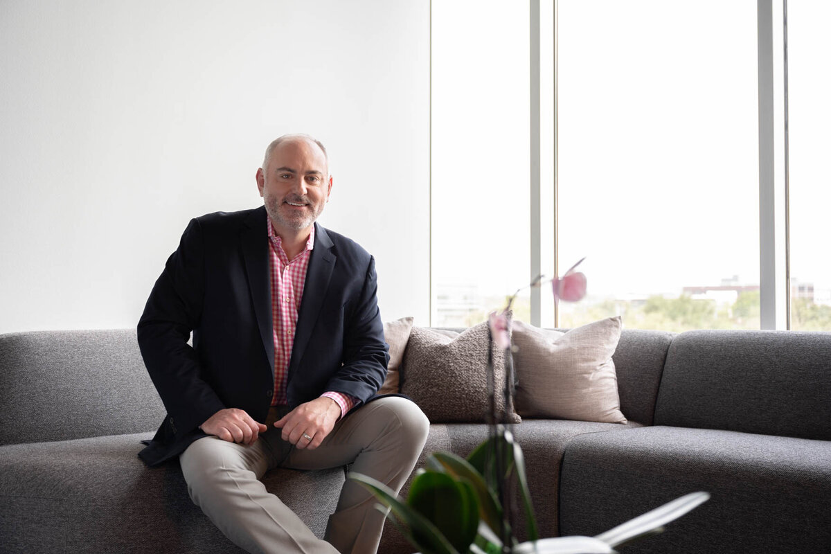 A man sitting on a couch smailing and next to him there is a big window with a view to town