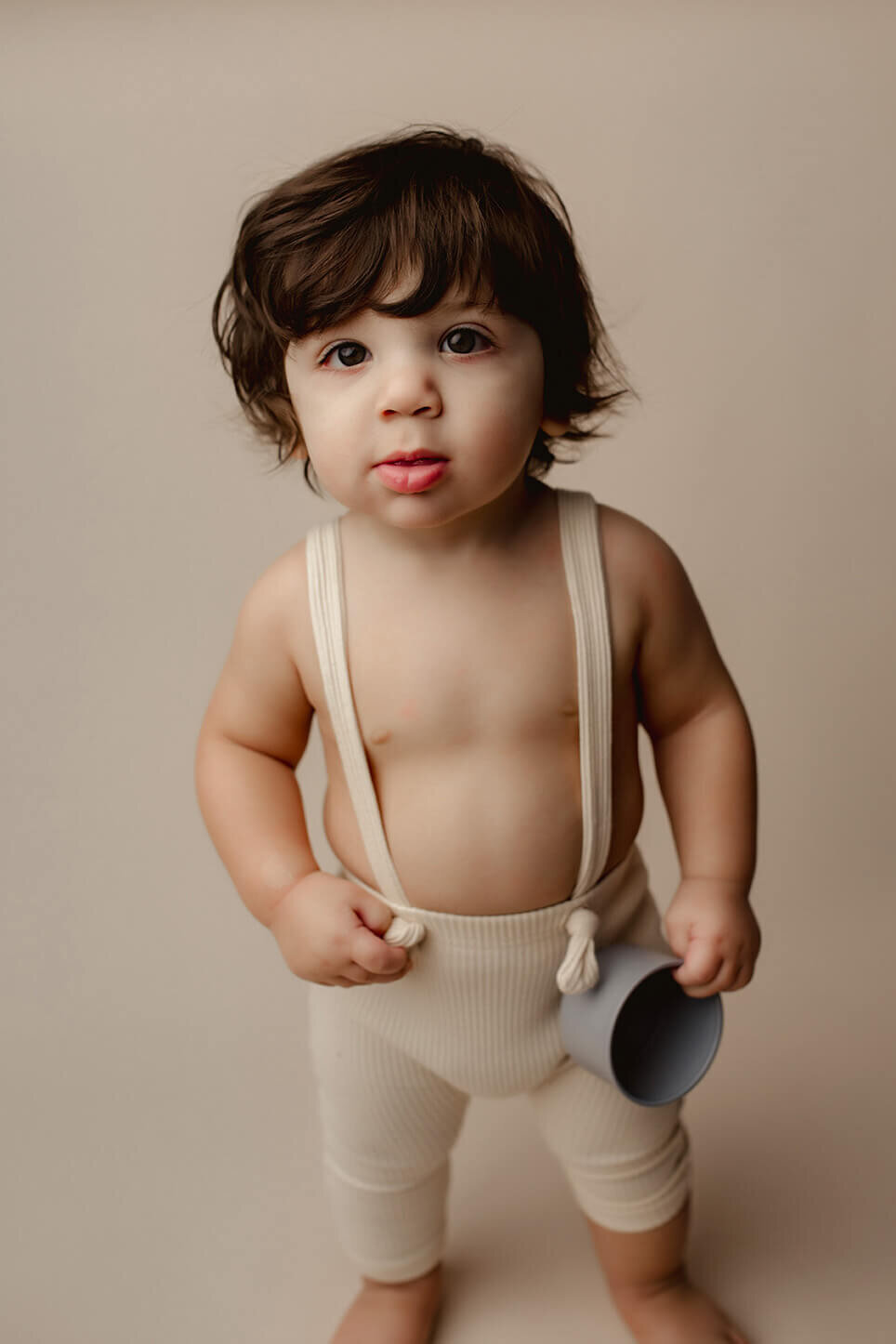 a 1 year old boy stands on a white backdrop with white suspenders on