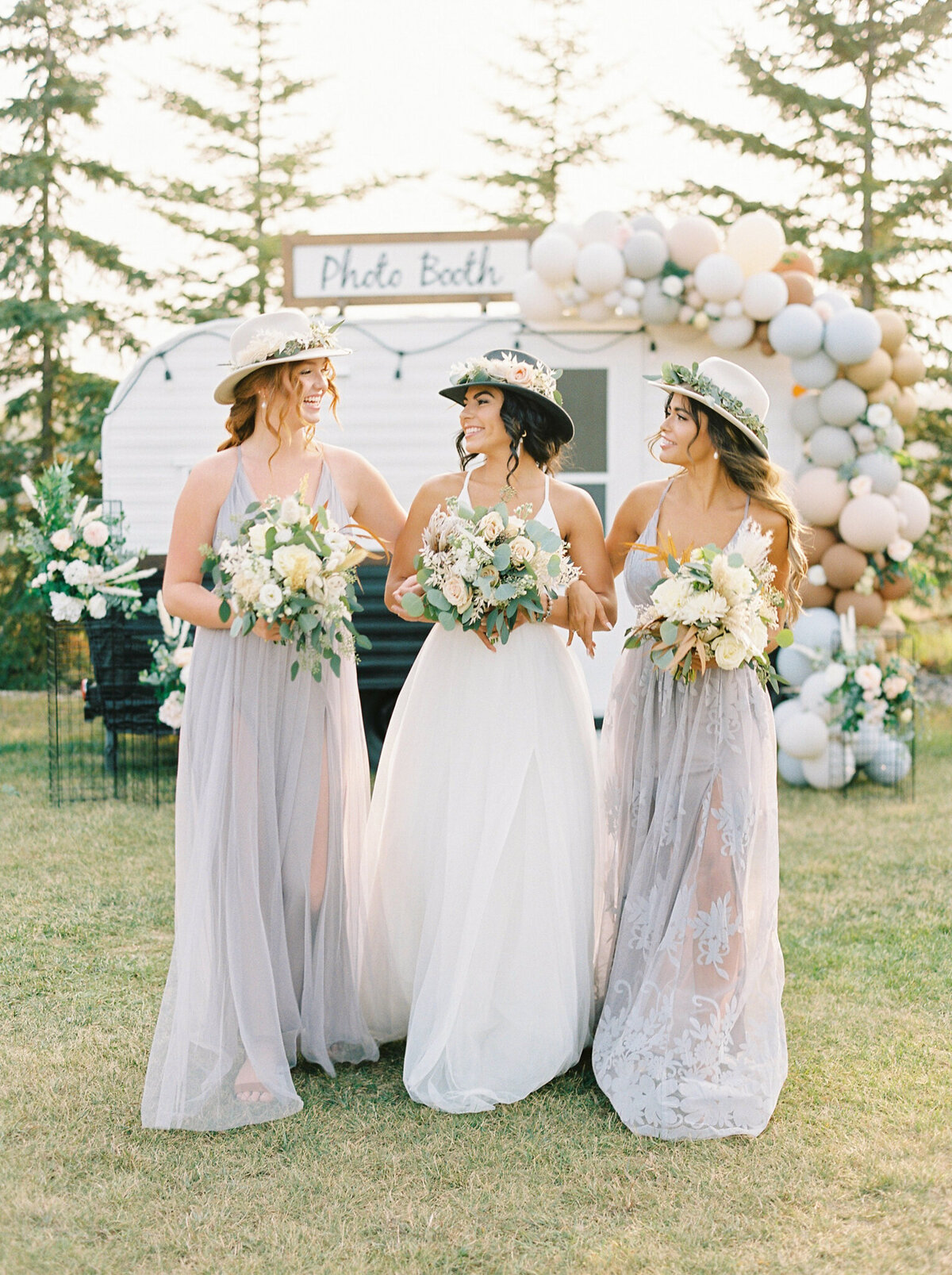 Gorgeous wedding inspiration, bride with bridesmaids wearing trendy wedding hats lined with florals and greenery in front of camper photo booth, captured by Justine Milton Photography, fine art  wedding photographer & videographer in Calgary Alberta. Featured on the Bronte Bride Vendor Guide.