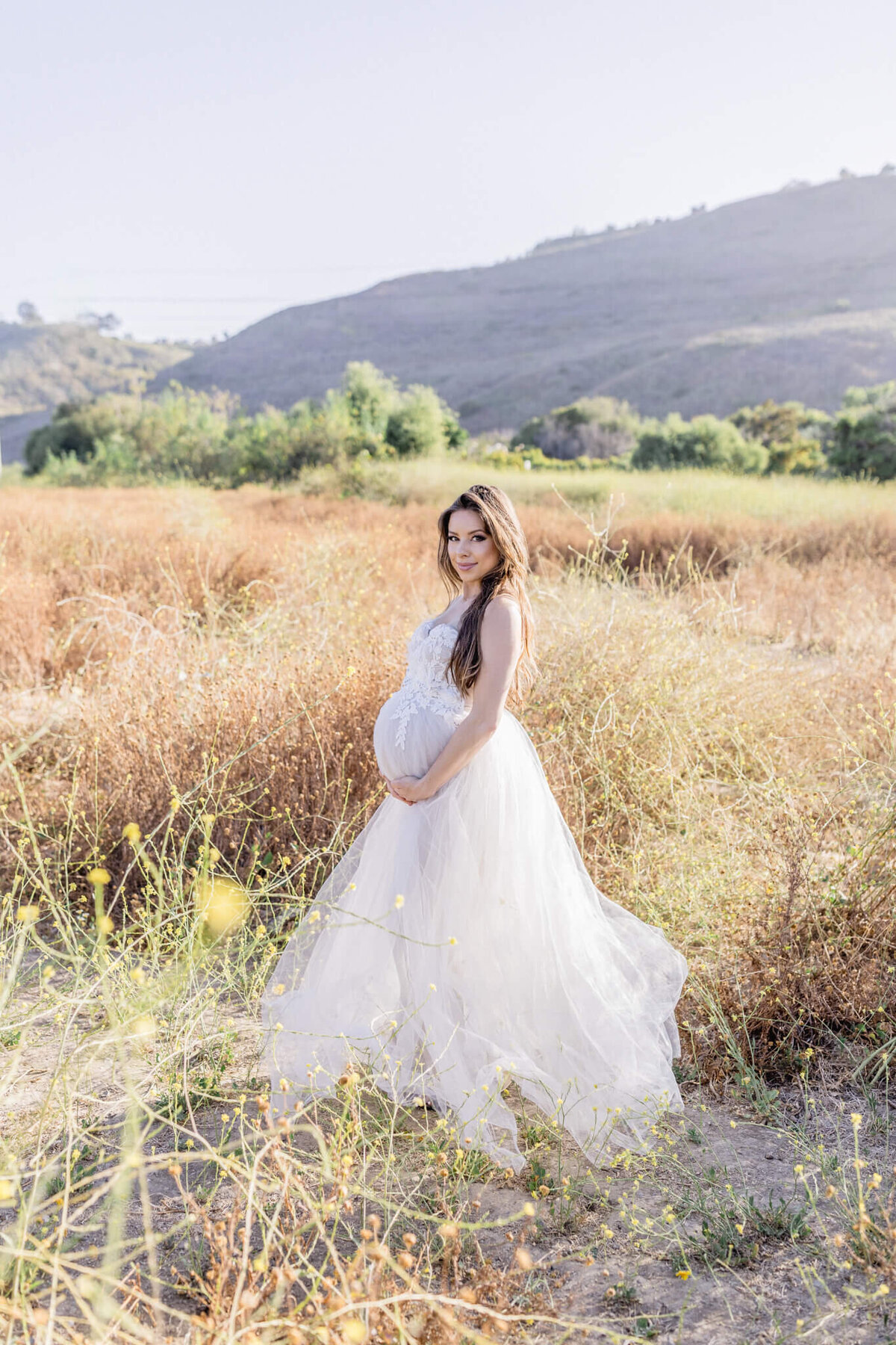 A pregnant woman in a flowing white dress stands in a sunlit field, with hills in the background. She cradles her stomach and looks at the camera, surrounded by dry grass and wildflowers. The sky is clear and bright.