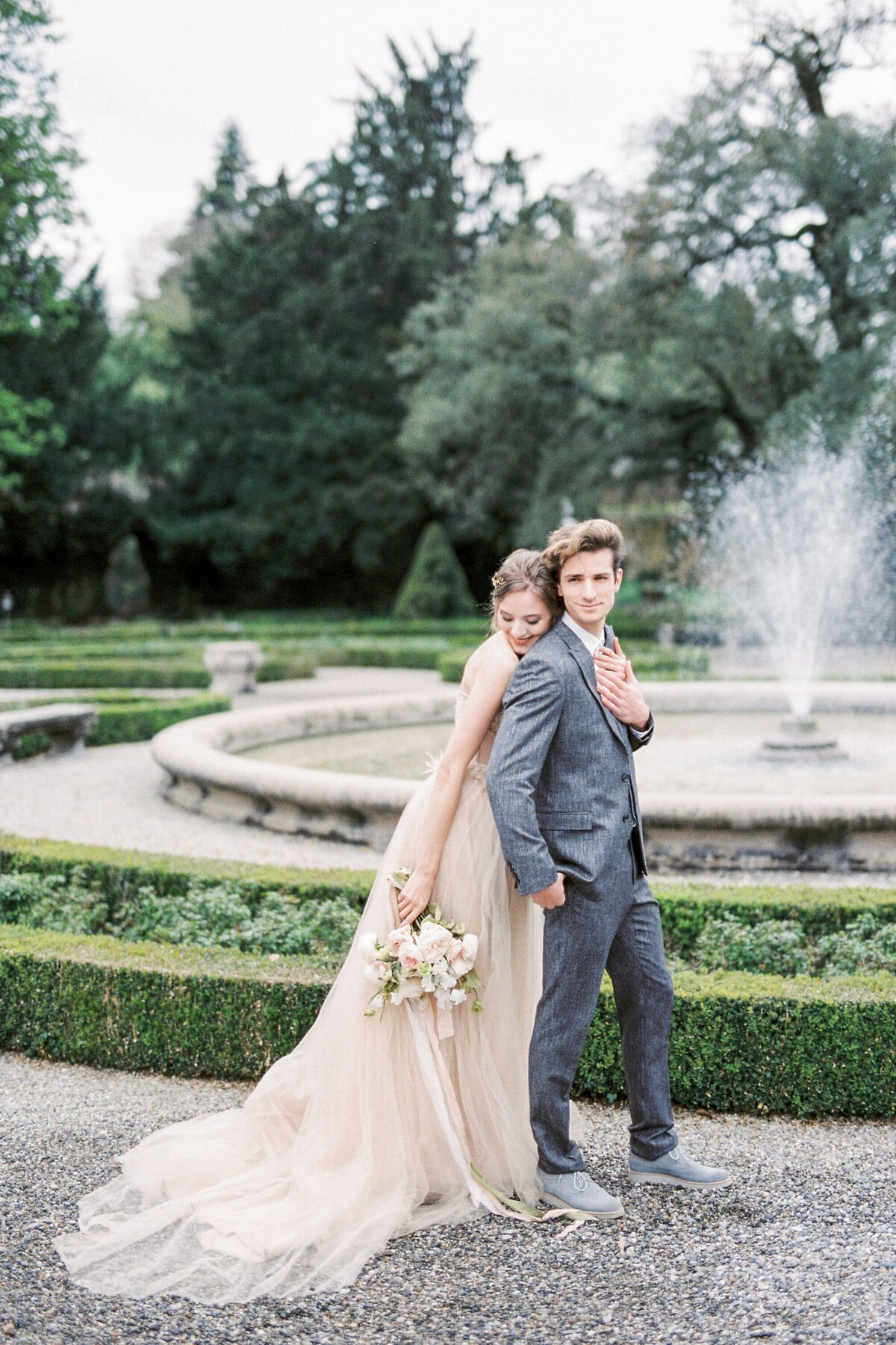 bride-with-bouquet-hugs-groom-from-near-fountain