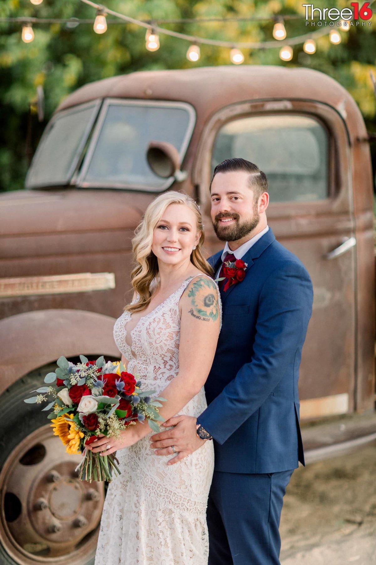 Groom holds his Bride's hips from behind as they pose in front of an old rustic-style truck behind them