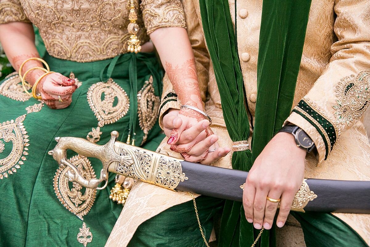 Sheikh groom wearing a green and gold sherwani holds a black sword with gold filigree detail sits next to a Hindu bride wearing a gold and green saree holding her hand.