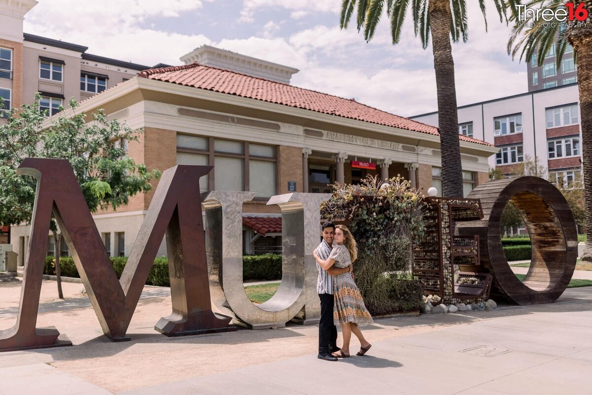 Engaged couple cuddle up to each other during an engagement photo session outside in front of the artistic lettering that spells Muzeo in front of the Anaheim Public Library