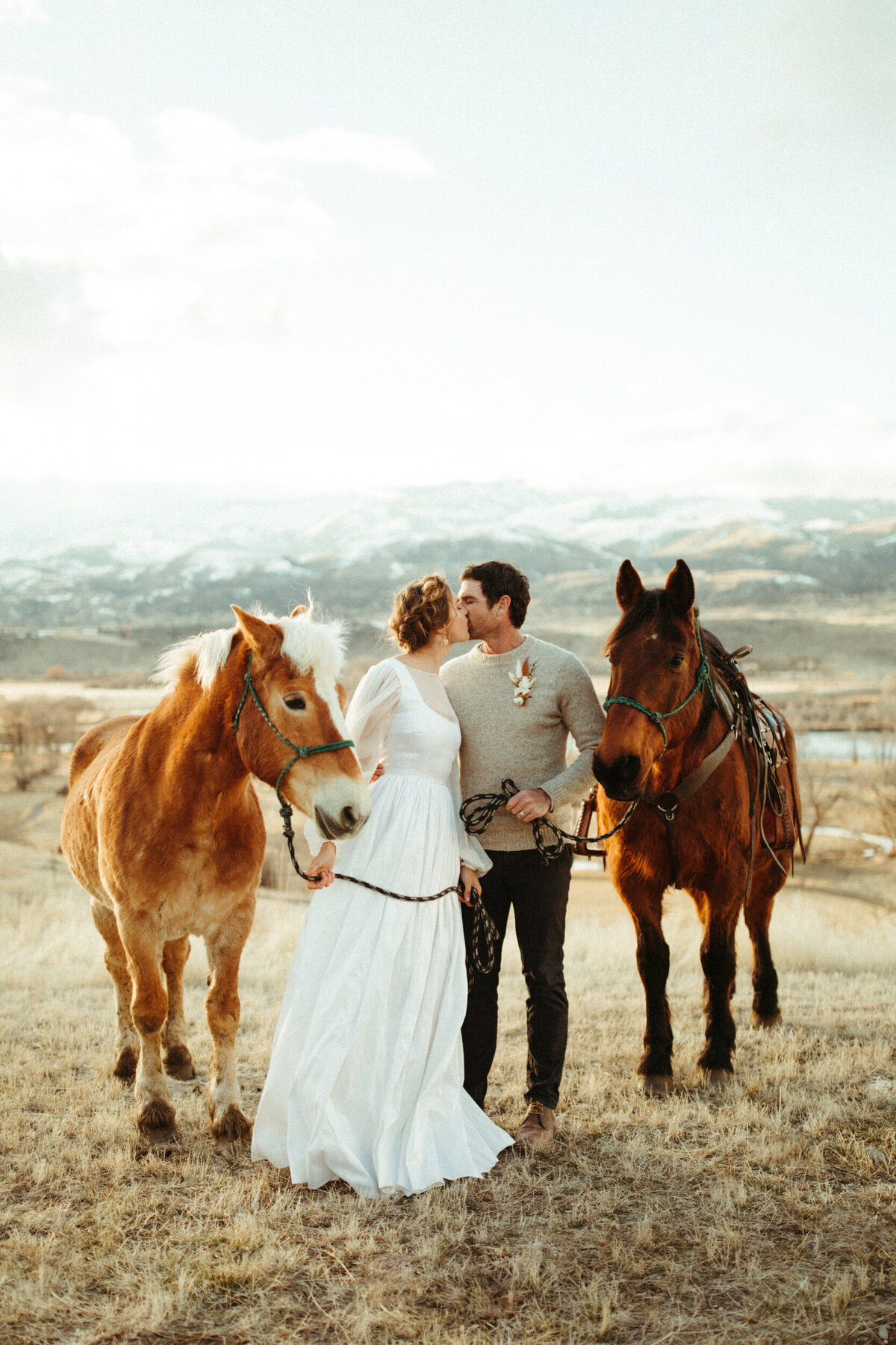 Bride and groom kissing and standing between two horses with snowcapped mountains behind them