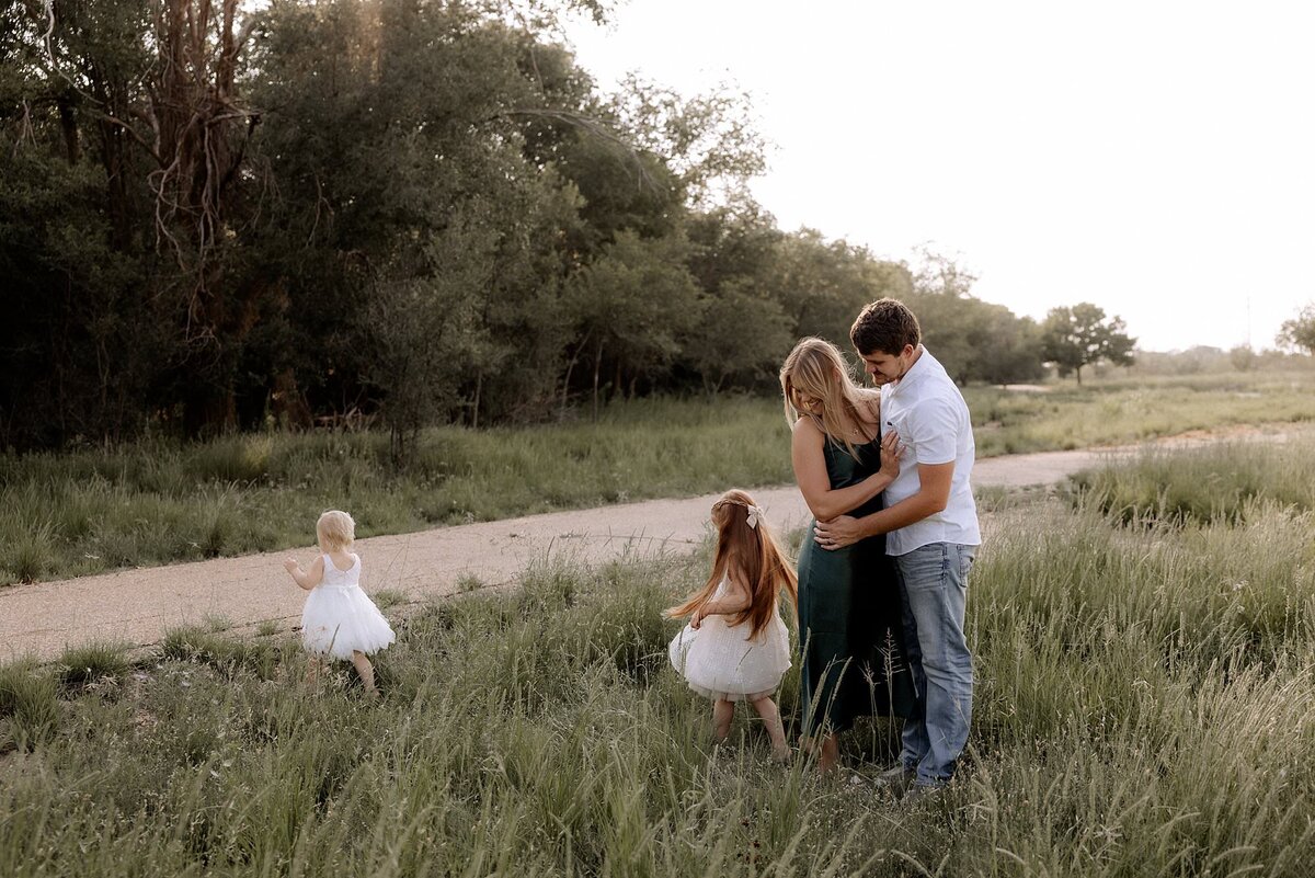 Father and Mother embracing while watching their daughters play in a filed of lush green grass.