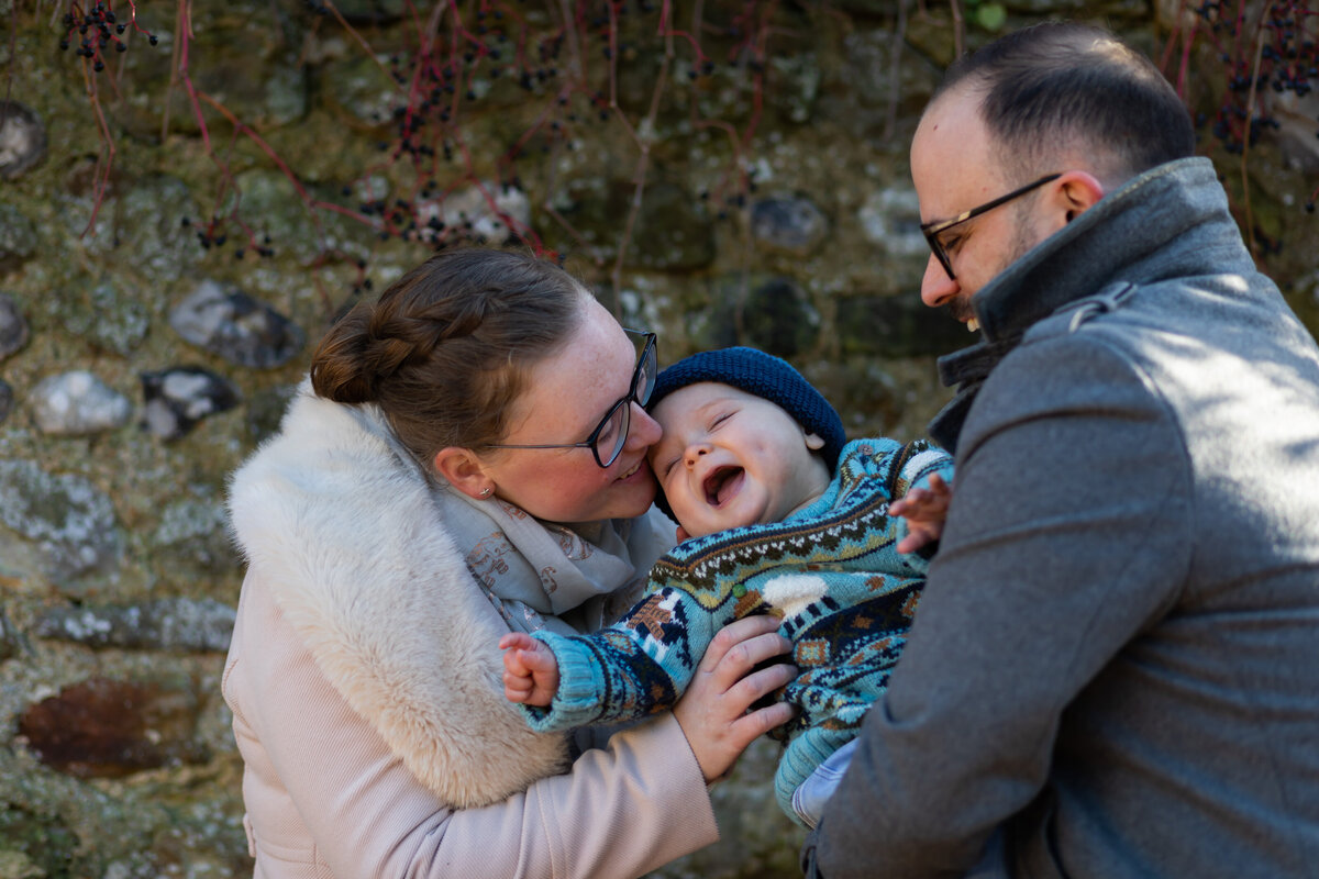 Family of three mum tickles toddler while dad holds him everyone is smiling