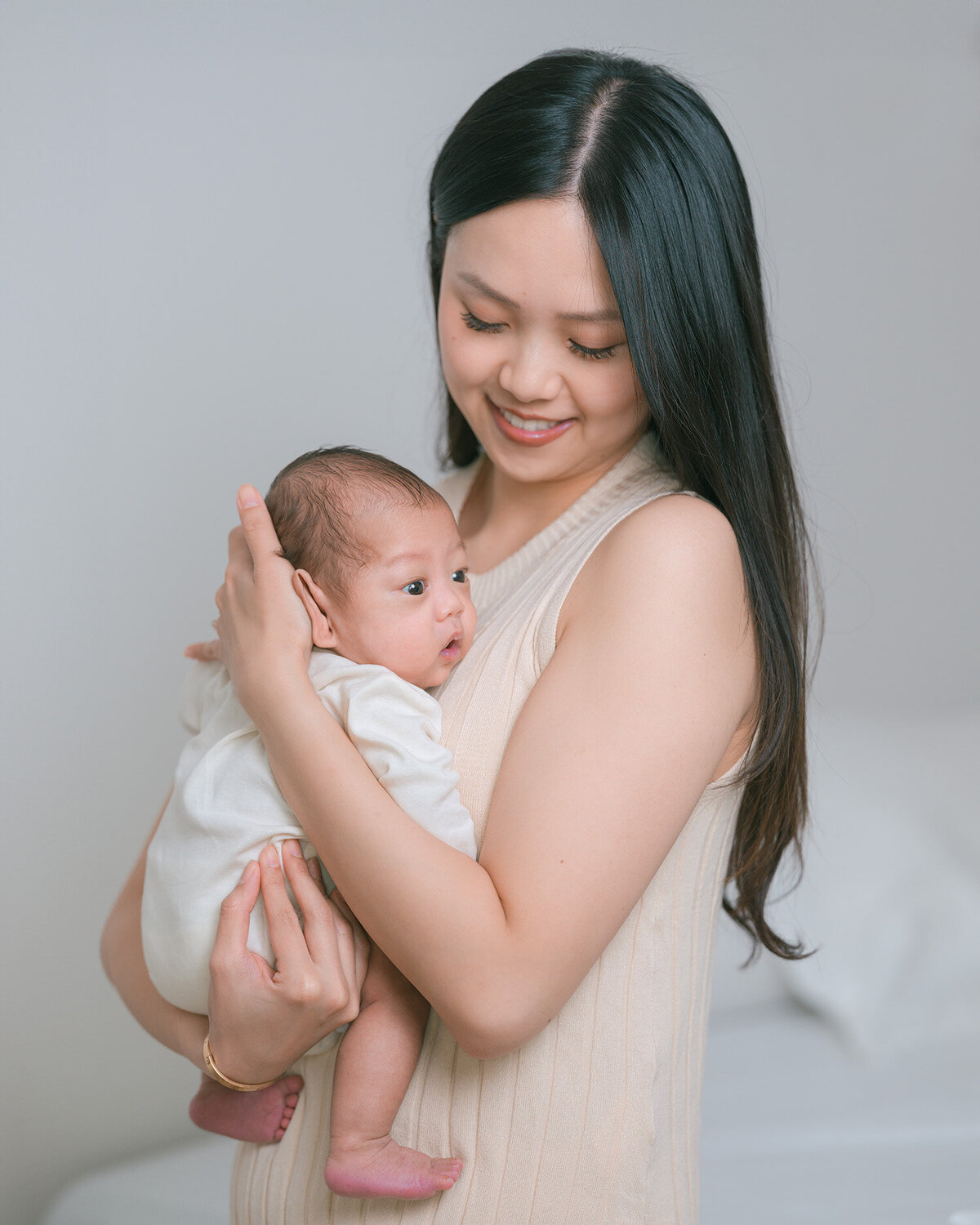 Young Asian mother wearing beige smiling while holding newborn baby in white long-sleeve onesie, photographed in natural light by Menlo Park newborn photographer.