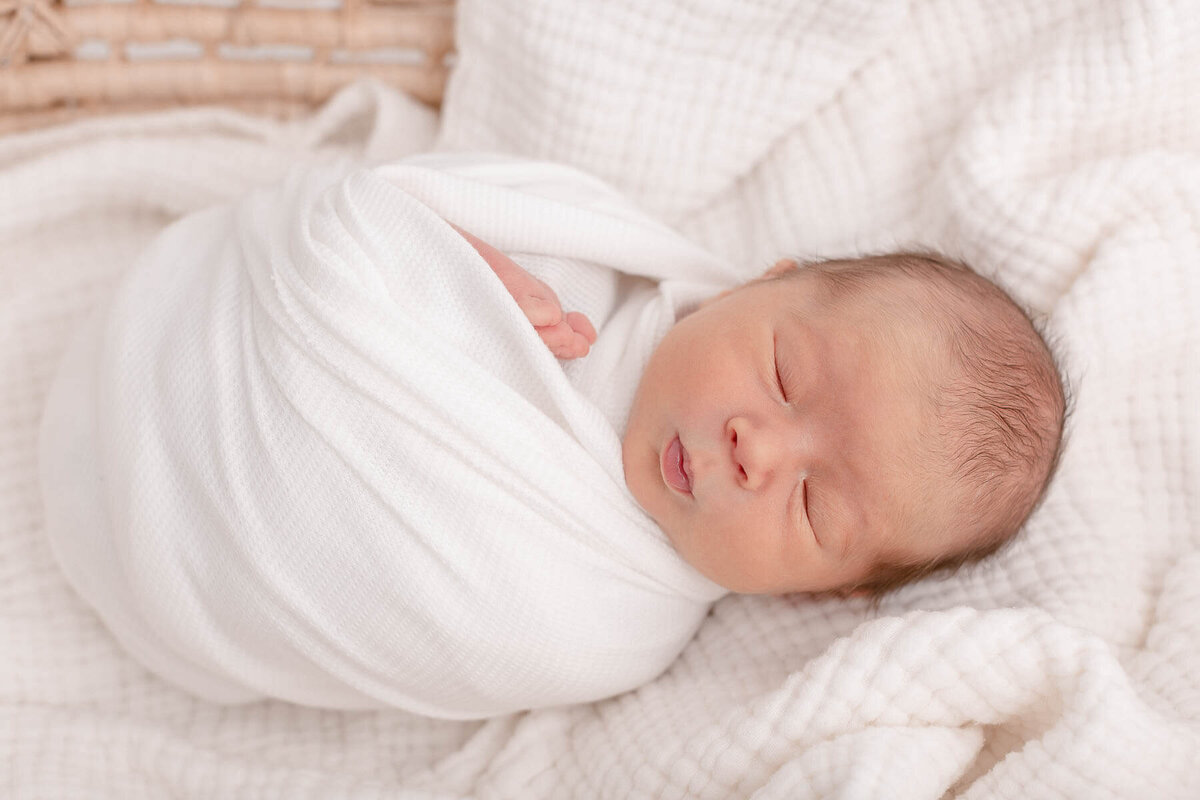 Sweet little newborn baby wrapped in white and laying on top of some white textured blankets inside of a moses basket.