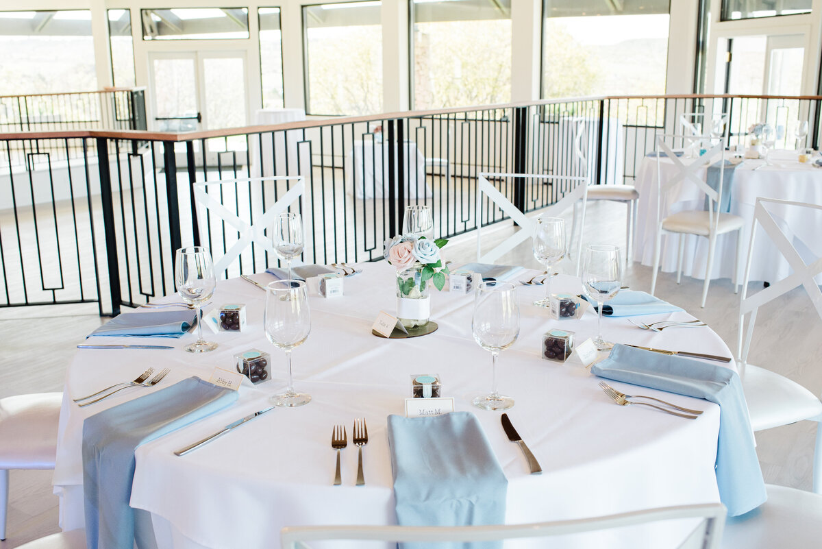 A photo of a reception table from a wedding at The Oaks at Plum Creek set up with light blue napkins, silverware, and small flower center piece