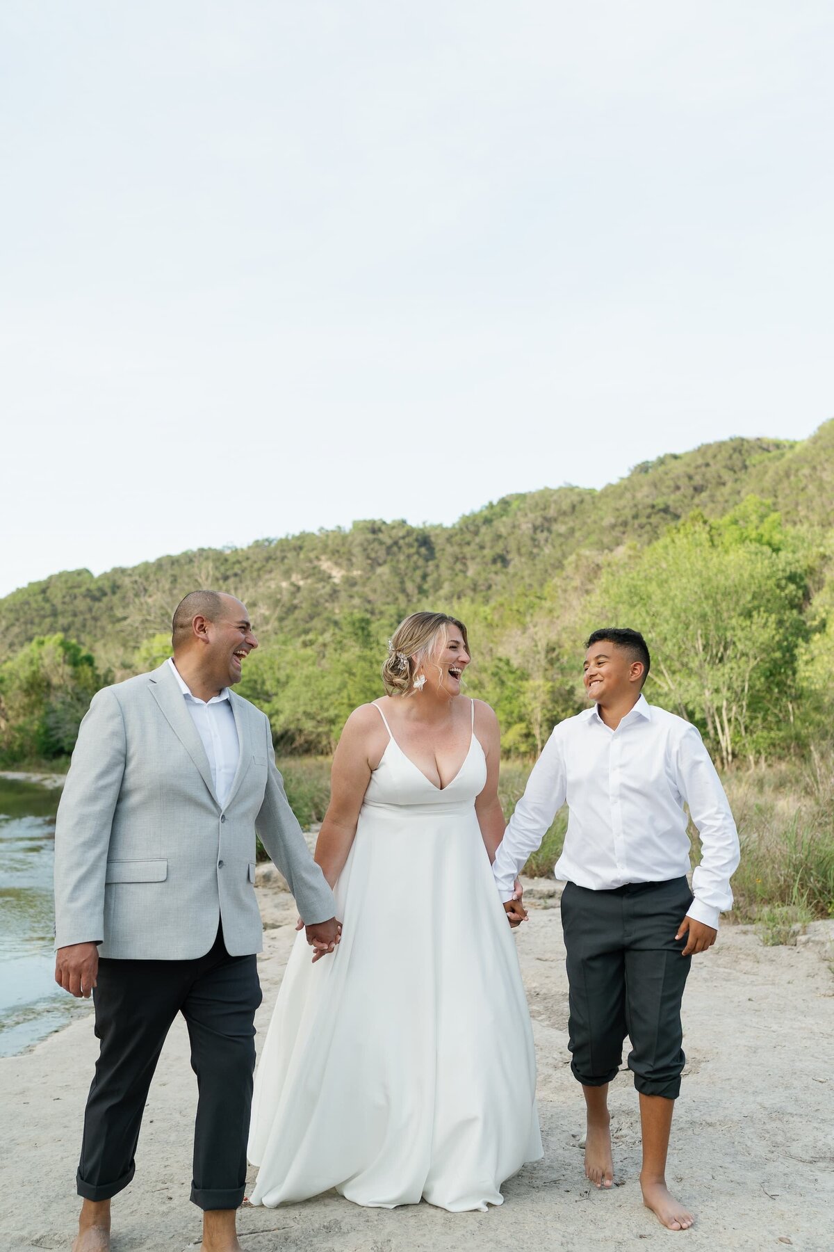 Jen, Miguel, and Deacon walking barefoot and laughing by the river during Austin elopement