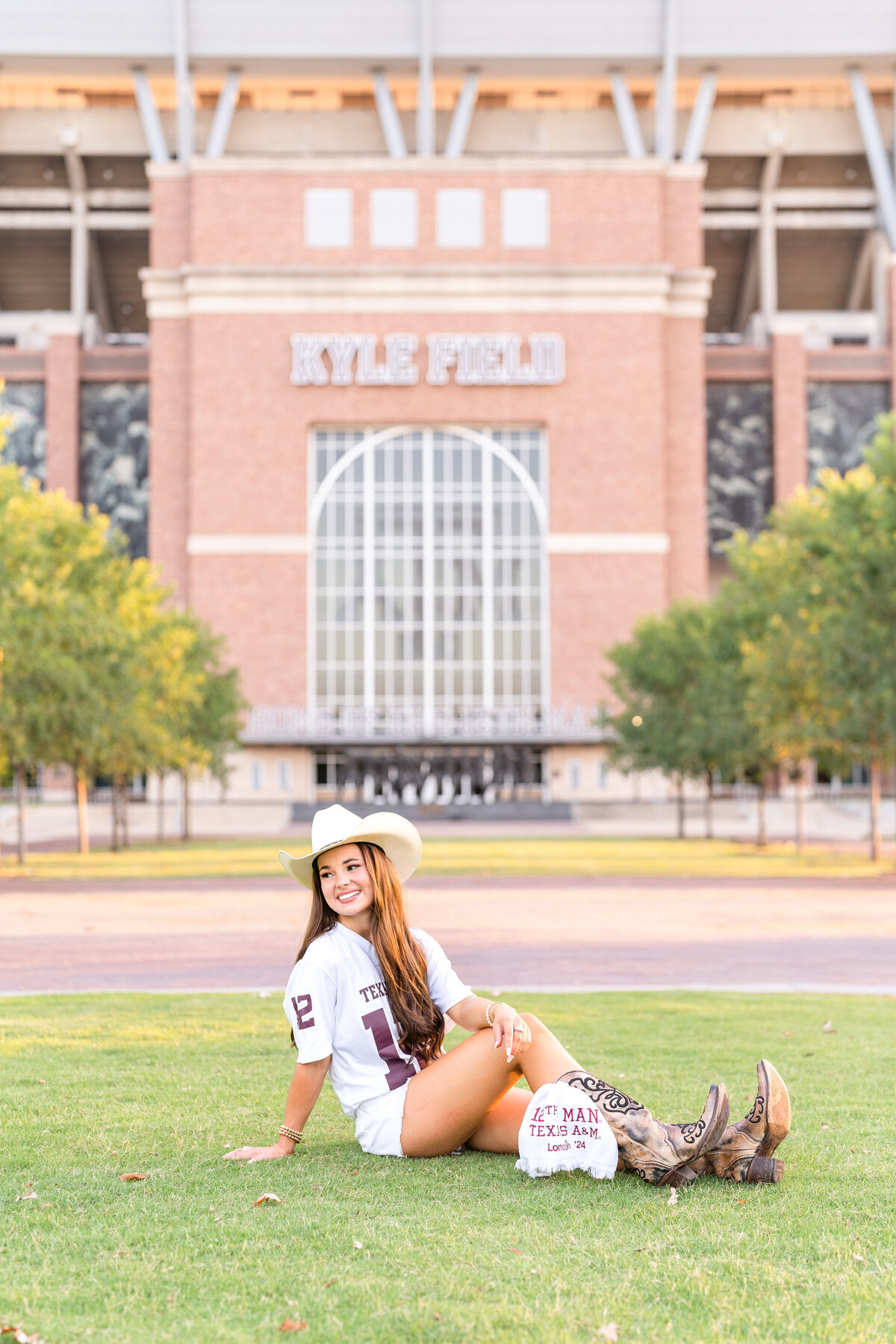 Texas A&M senior girl sitting in grass and looking over shoulder while wearing cowboy hat, boots and Aggie white jersey with Kyle Field in background in Aggie Park