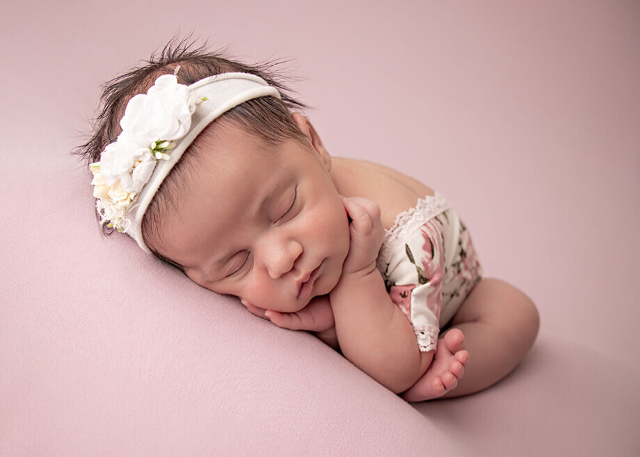 Newborn baby girl  in the timber pose on a soft pink beanbag backdrop.  Wearing a white floral bodysuit and a matching floral headband