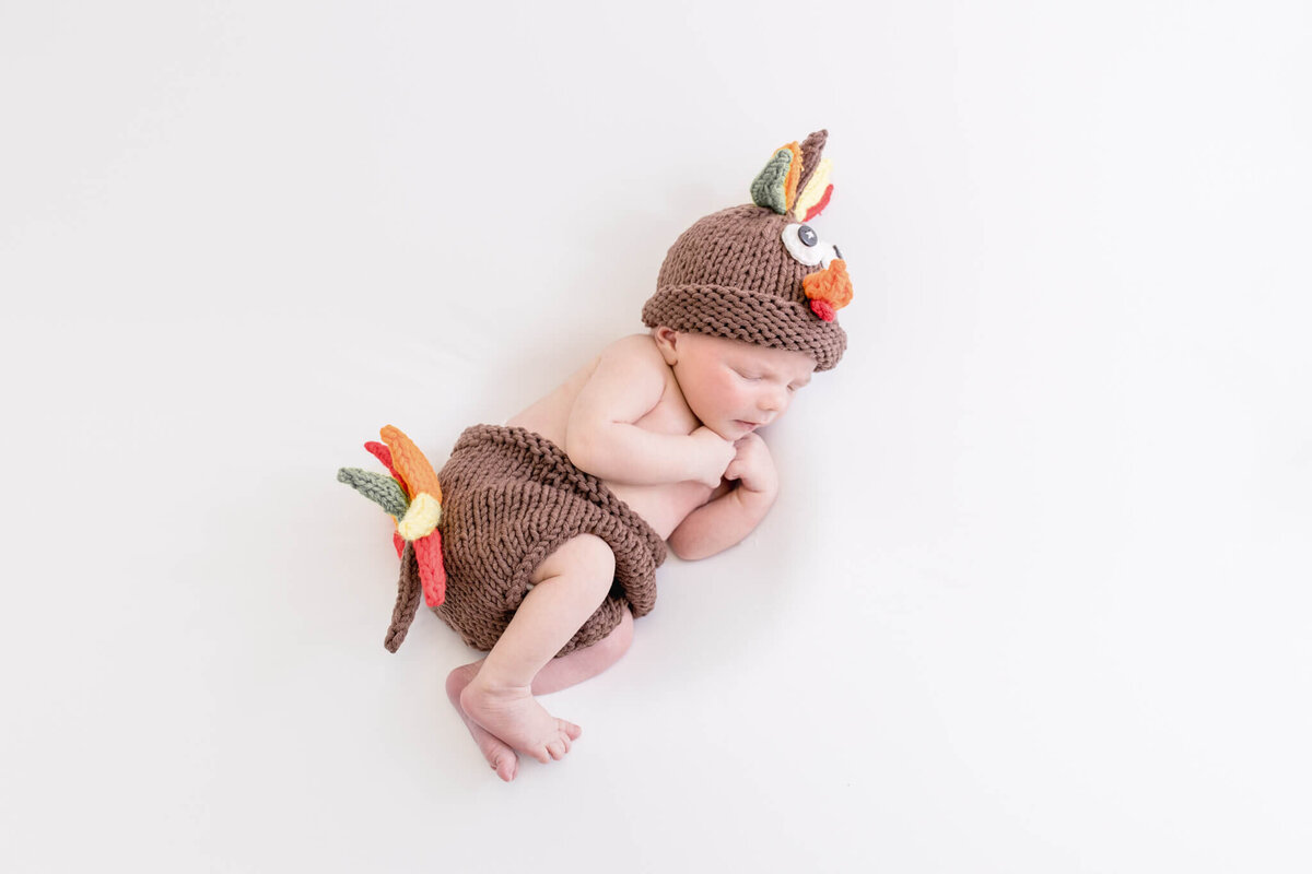 A baby is peacefully sleeping, dressed in a knitted brown turkey costume with colorful feathers on the hat and shorts. The background is plain white.