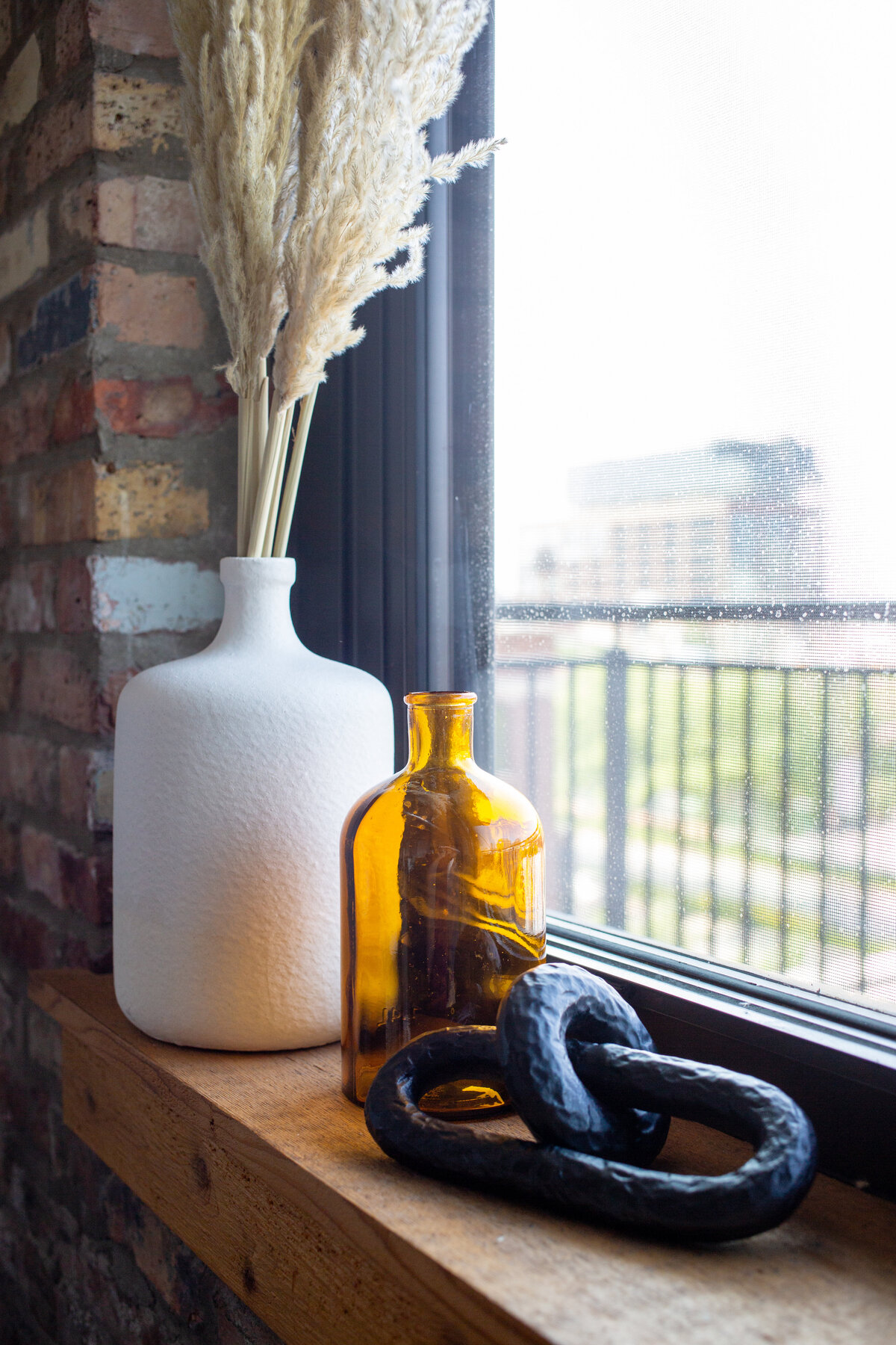 Brick wall with wooden window sill with white ceramic vase filled with pampas grass, amber glass vase and black chain object