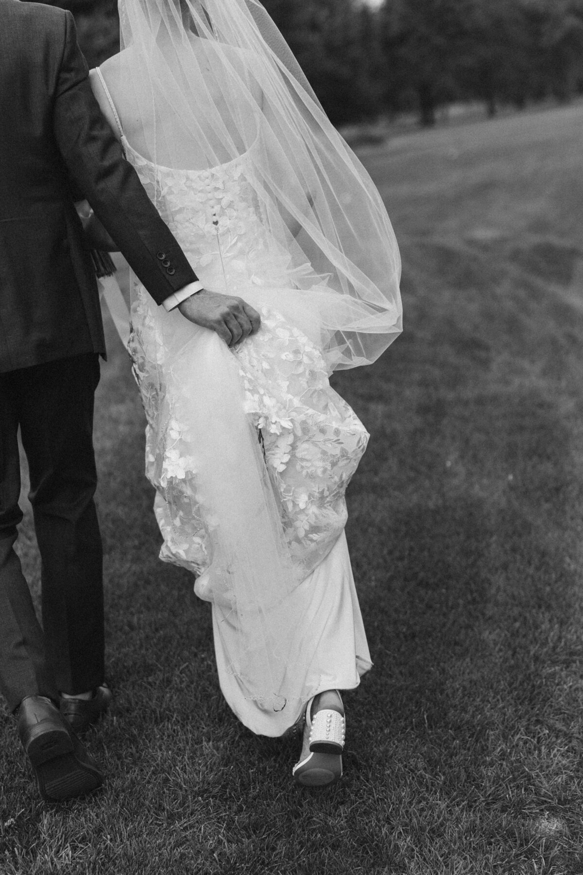 Groom holding bride’s lace wedding dress as they walk together across a grassy field, showing her pearl-studded heels and flowing veil.