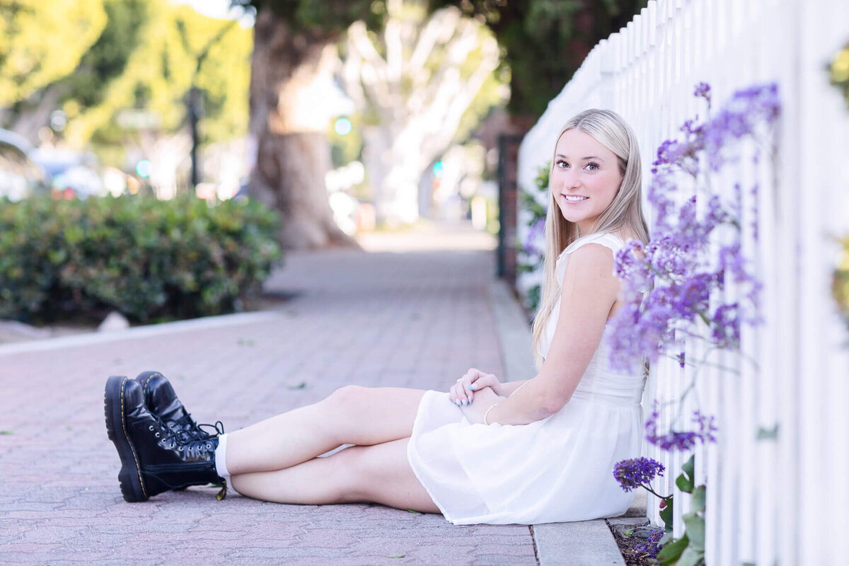 A woman with long blonde hair is sitting on a sidewalk next to a white fence adorned with purple flowers. She wears a white dress and black boots, and smiles while looking at the camera. The background shows trees and a sunny street.