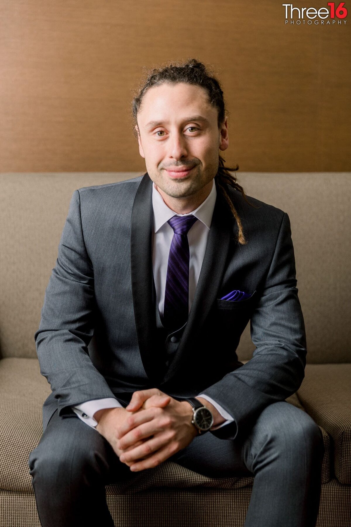 Groom sits on the edge of couch with his hands folded as he poses for photos prior to his wedding ceremony