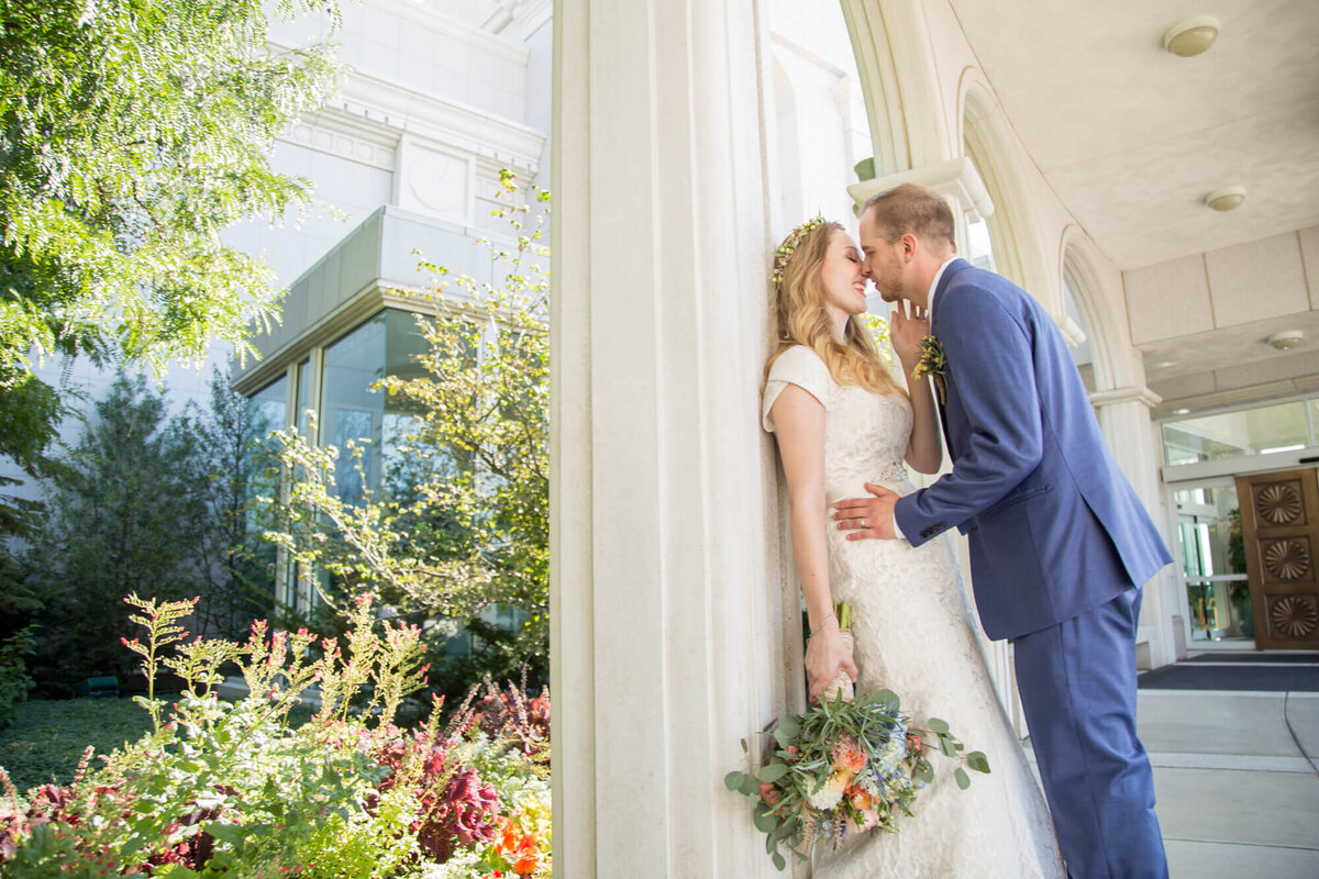 bride in a classic lace dress against a stone pillar with groom leaning in for a kiss