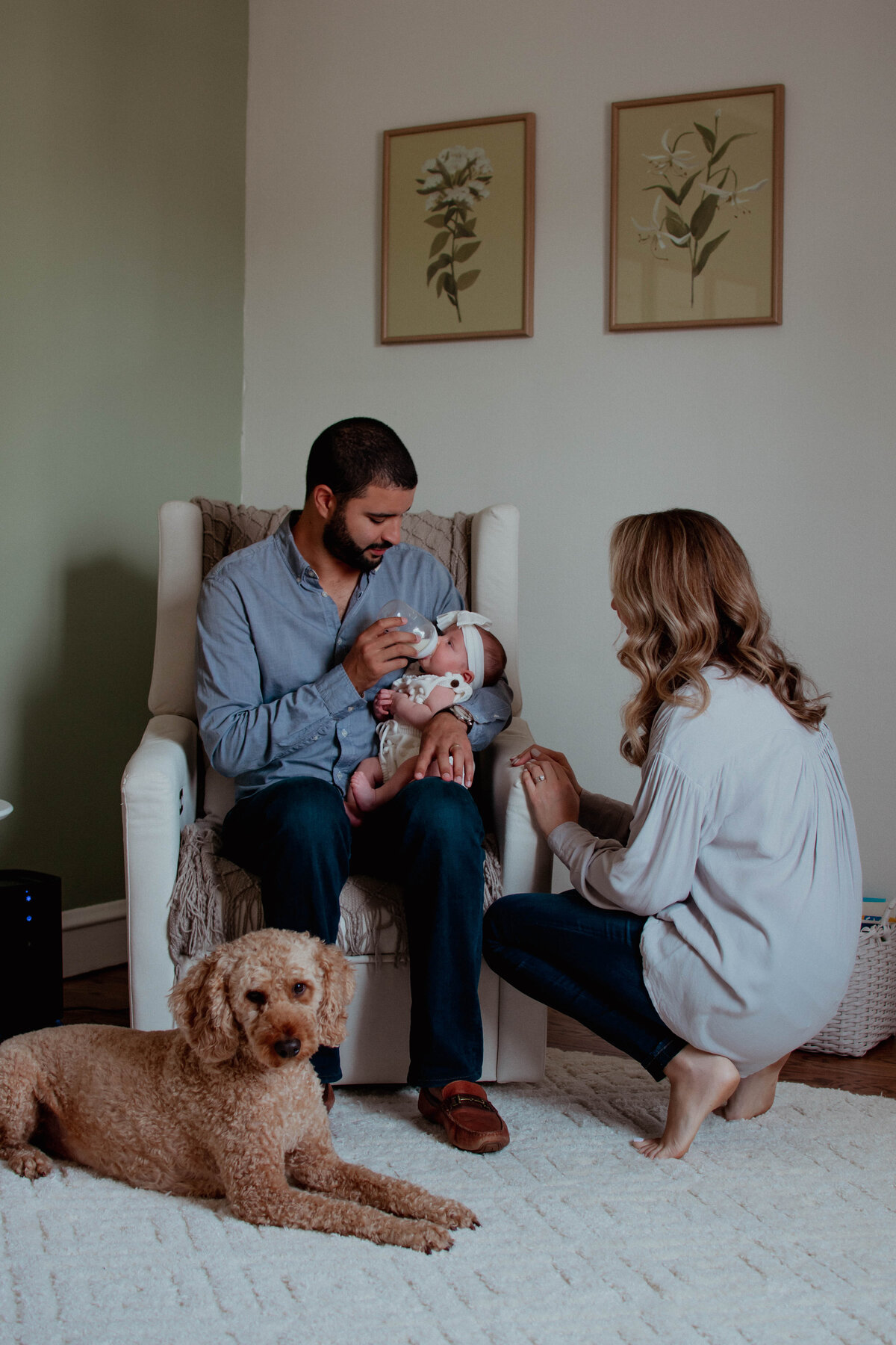 Mom admiring dad feeding their baby girl while the dog naps on the nursery floor during a newborn photoshoot in Chicago, IL