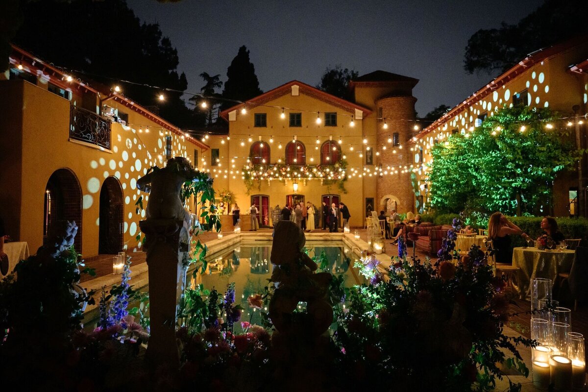 A night photo of a Whimsical Paramour Estate Wedding. You can see the Paramour Mansion Pool and estate illuminated. Garden flowers and ivy climb the statues around the pool.