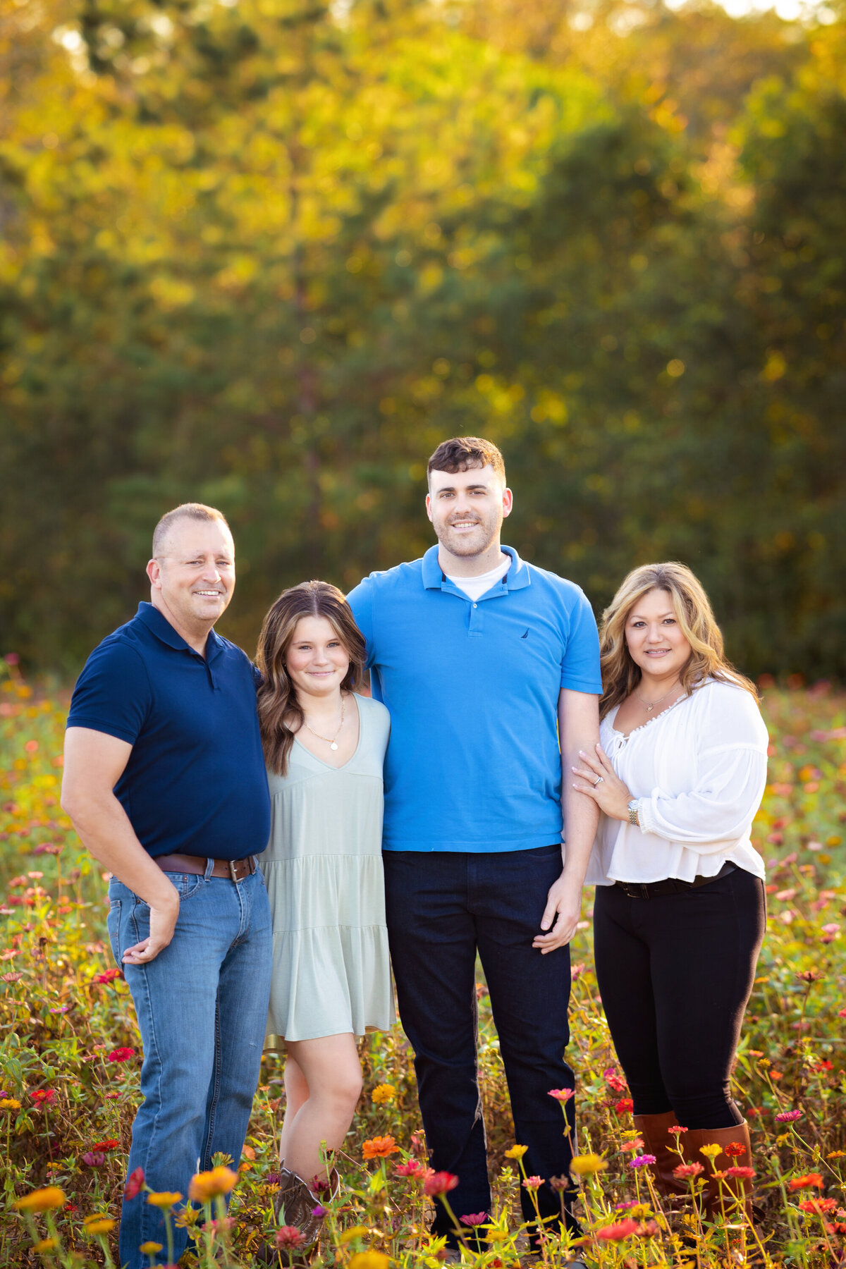 Family with teenage girl and adult sun standing in the zinnia field.  Dad is wearing a navy shirt and jeans.  Mom is wearing black leggings and a white shirt.  Teenage girl is wearing a pale mint dress and boots.  Adult son is wearing black jeans and a blue polo shirt.