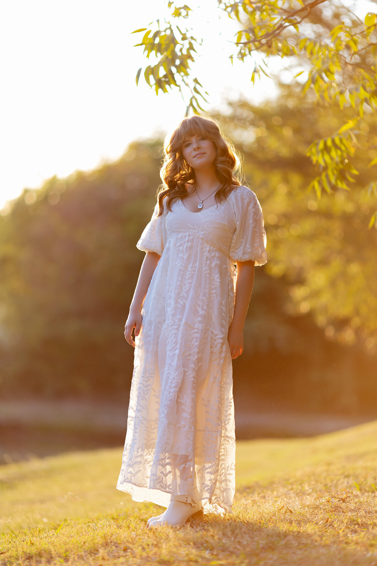 Dallas senior photography of a young woman in a white dress, standing in a golden sunset with soft bokeh in the background.