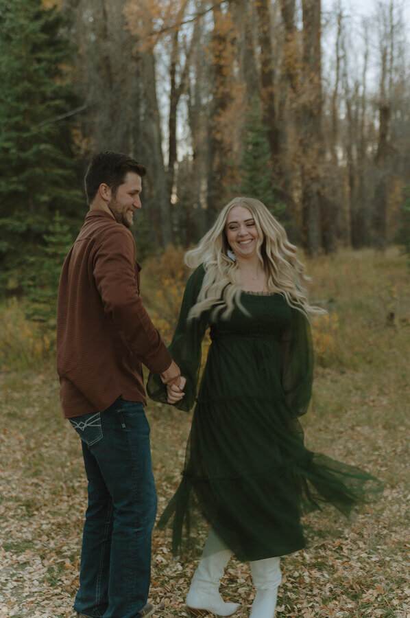 A man spins his fiancé in a forest. Her dark green dress twirls with motion.