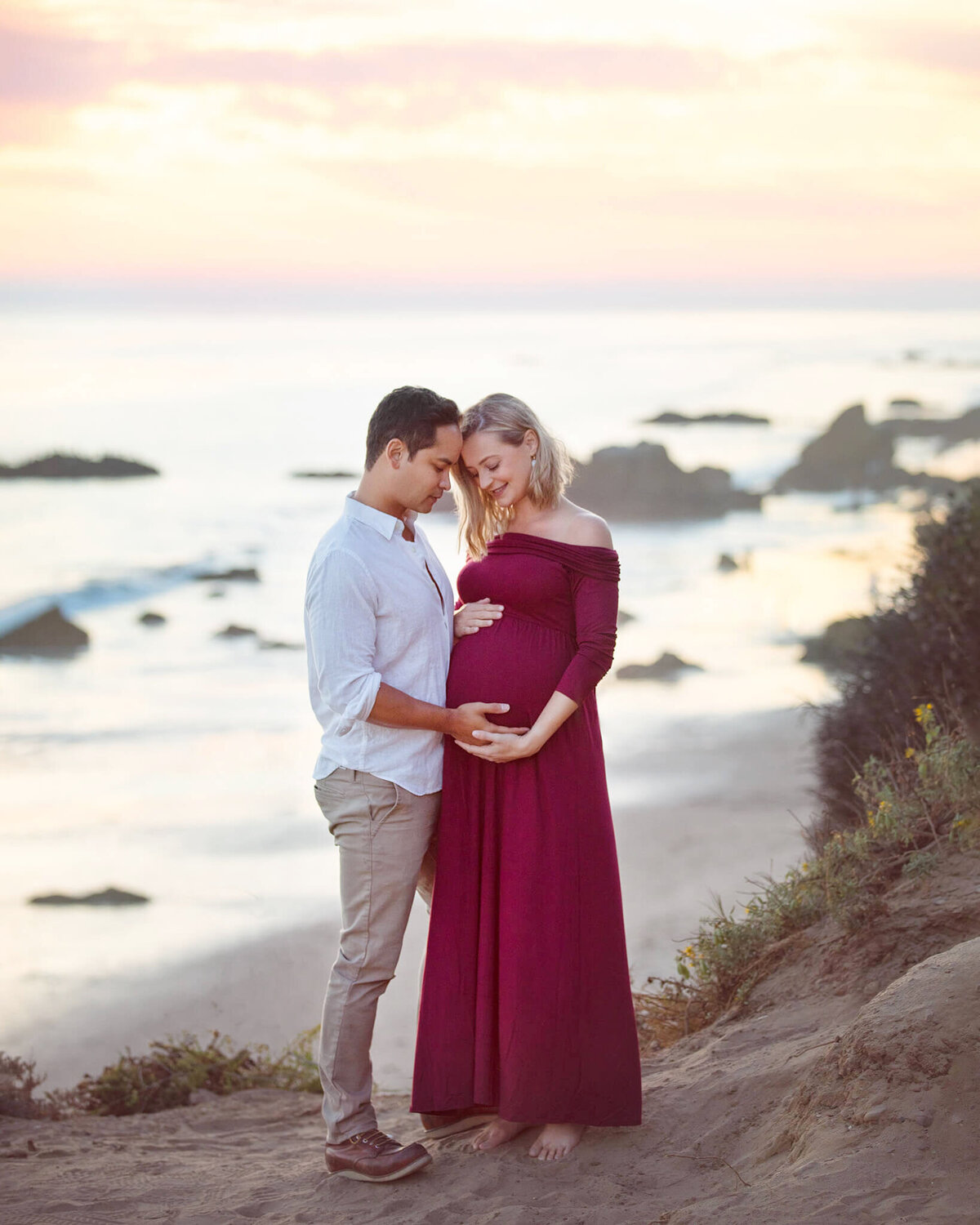 Mom and dad to be with heads together looking at baby bump with mom wearing a red dress at El Matador Beach in Malibu