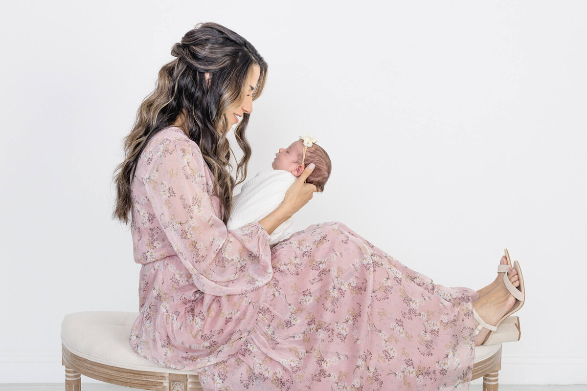 A woman with long hair in a pink floral dress is sitting on a bench, gently holding and looking at a newborn baby wrapped in white. The baby is wearing a small headband. They are set against a plain white background.