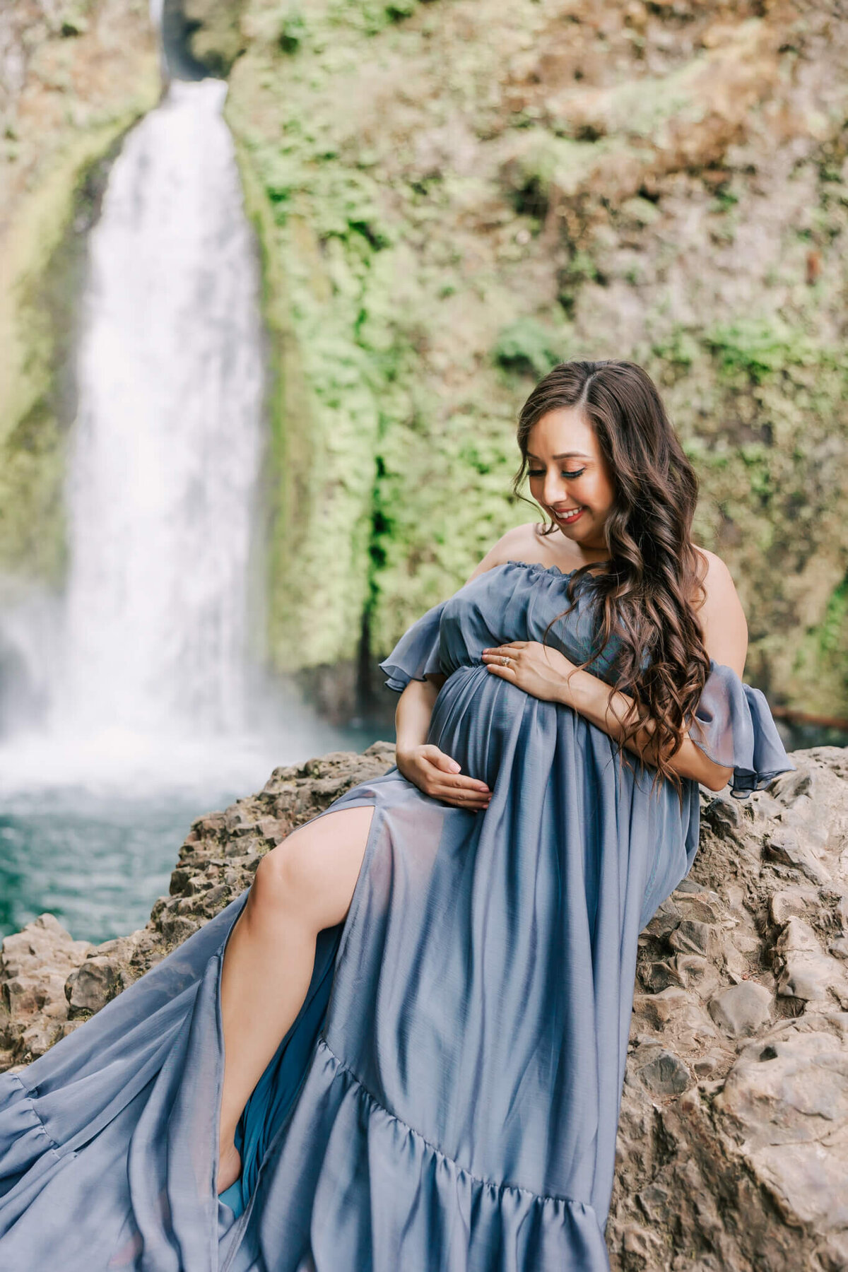 portrait of a woman in a blue dress with long brown hair smiling down at her pregnant belly with her hands on her stomach. There is a waterfall in the columbia river gorge behind her.